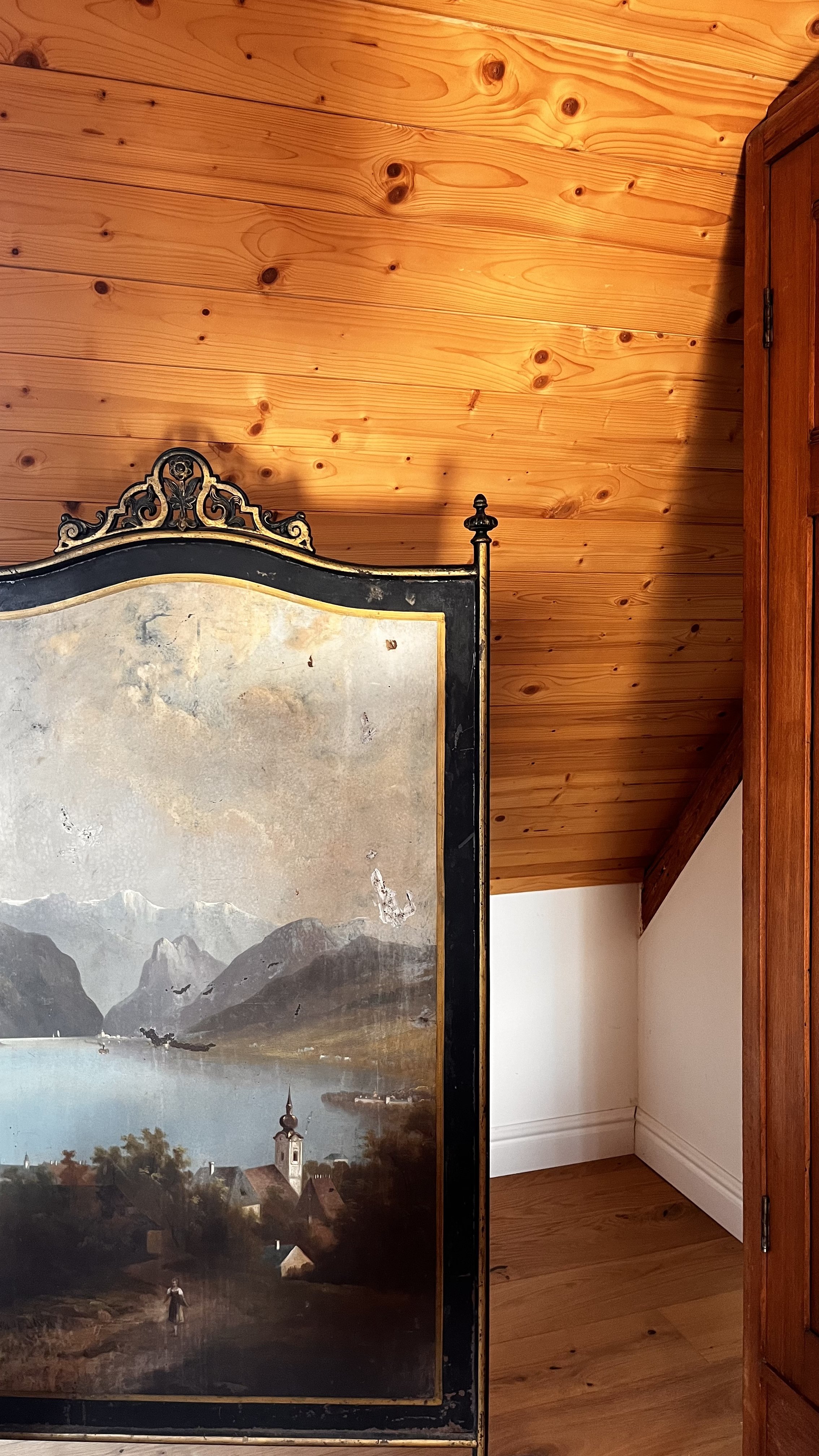 Part of an antique painting with a landscape scene, including mountains, water, a village, and a church, placed against a wood-paneled wall and ceiling.