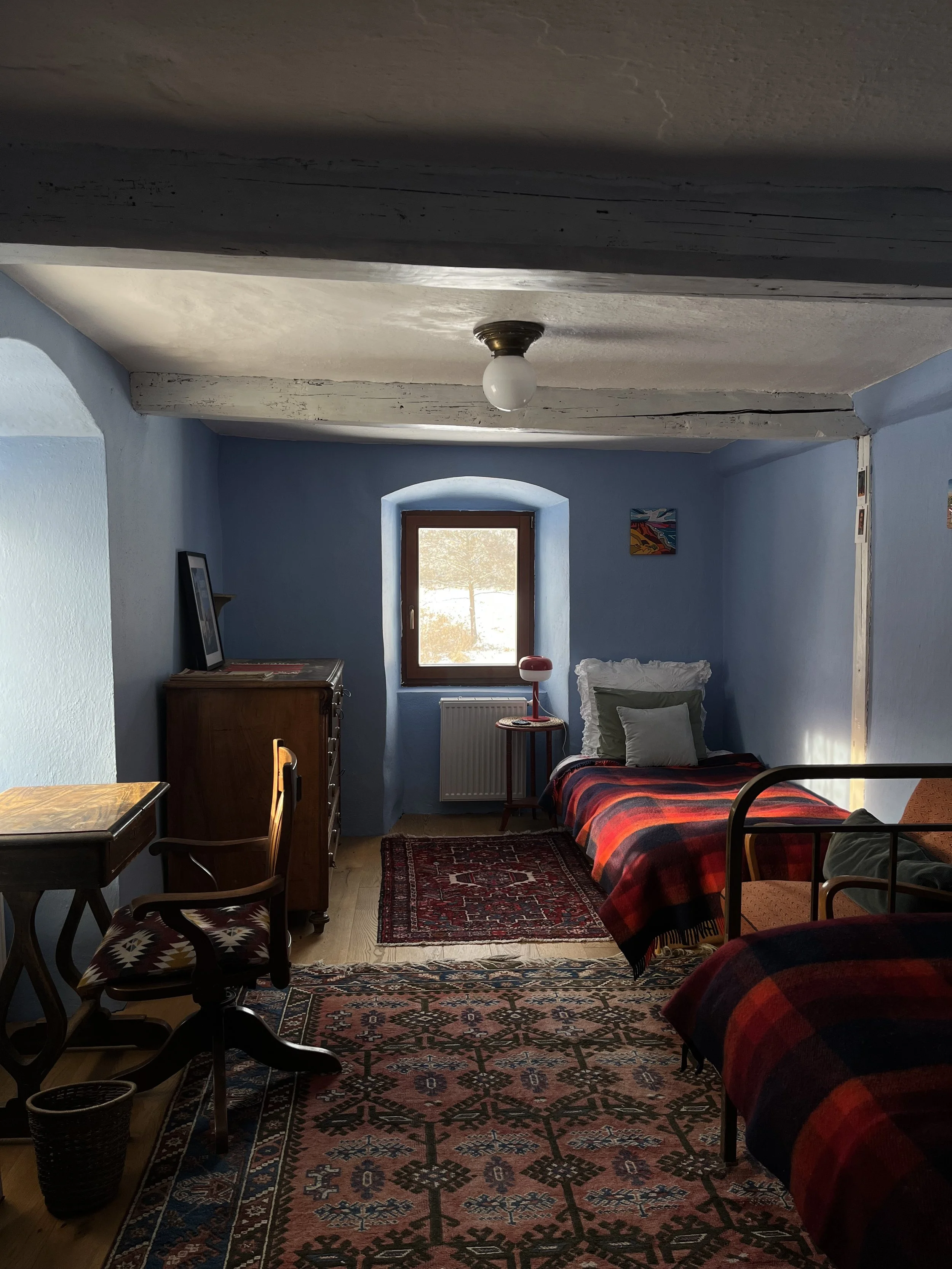A cozy bedroom with light blue walls, two single beds with red and black plaid blankets, a window with a view of snow outside, a small wooden table with a red lamp, a vintage dresser with a picture frame, a wooden desk with a chair, colorful rugs, and a painting on the wall.