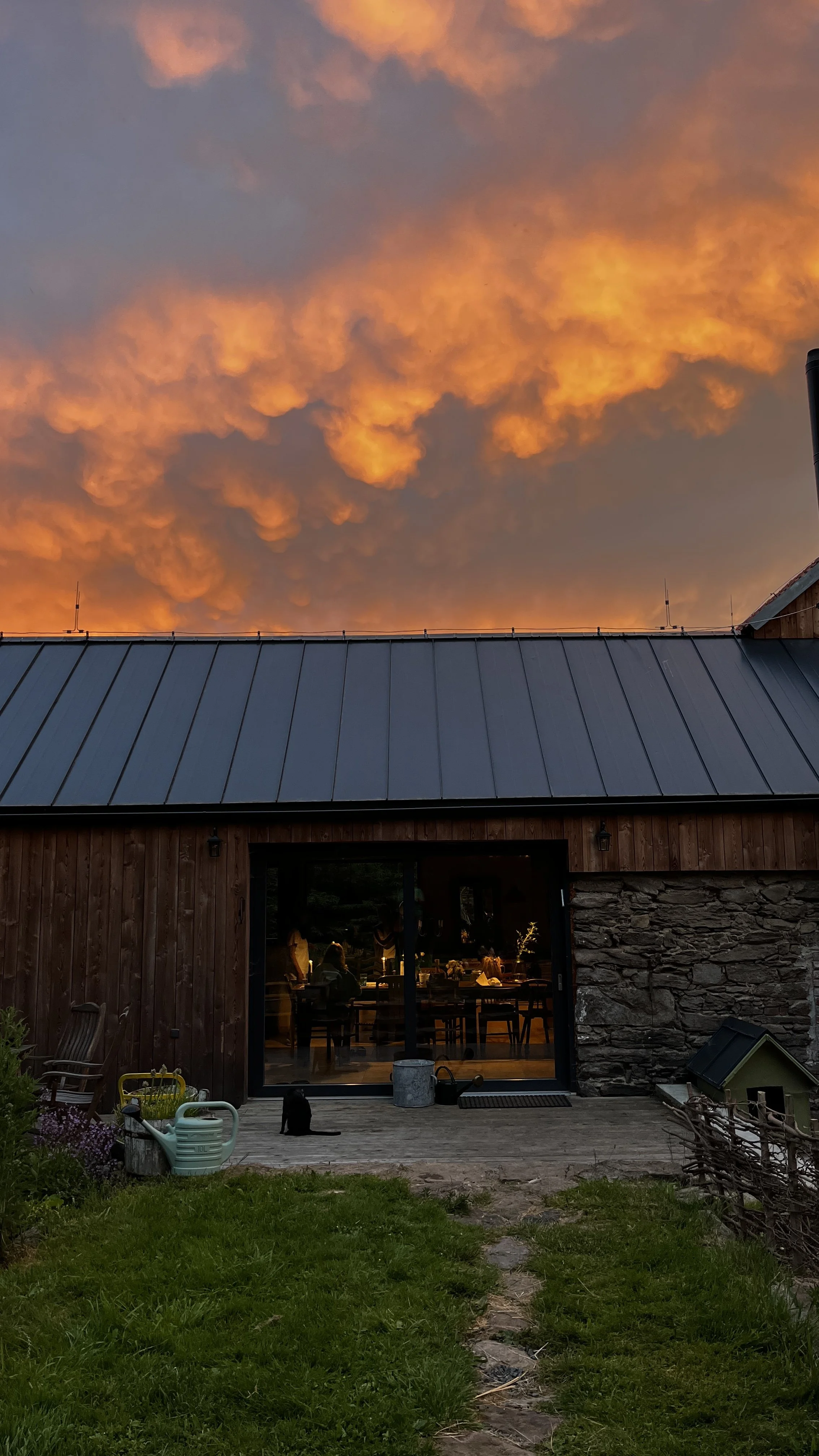A rural scene with a barn with a dark metal roof, large sliding glass door, and stone wall in Bohemia. Inside, people are sitting at tables. Outside, a black cat sits on the deck. The sky above shows vivid orange clouds at sunset.