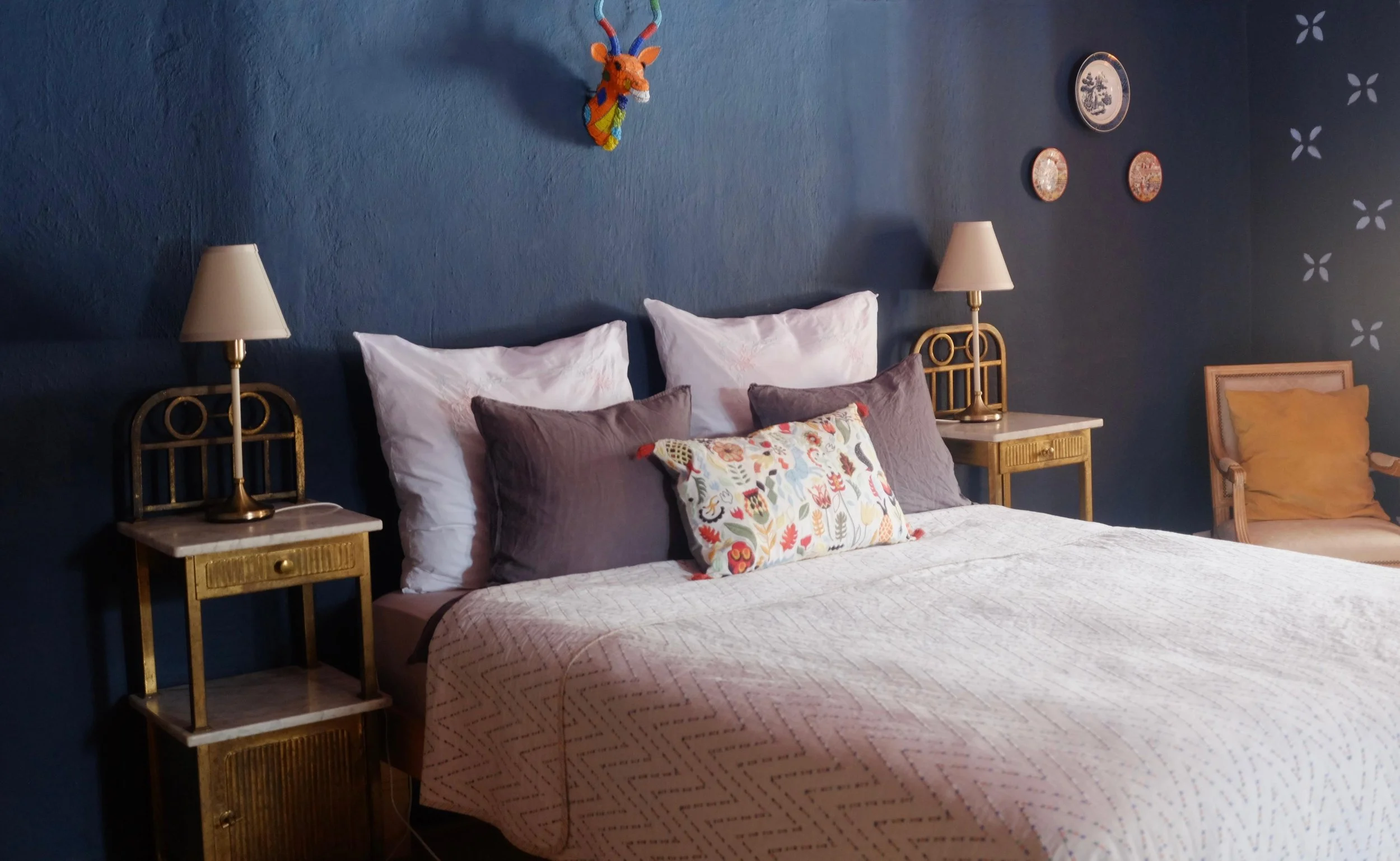 A bedroom with a dark blue wall, decorated with a colorful giraffe wall hanging and three small plates. There are two nightstands with lamps, a bed with white, purple, and patterned pillows, and a chair with a yellow pillow.