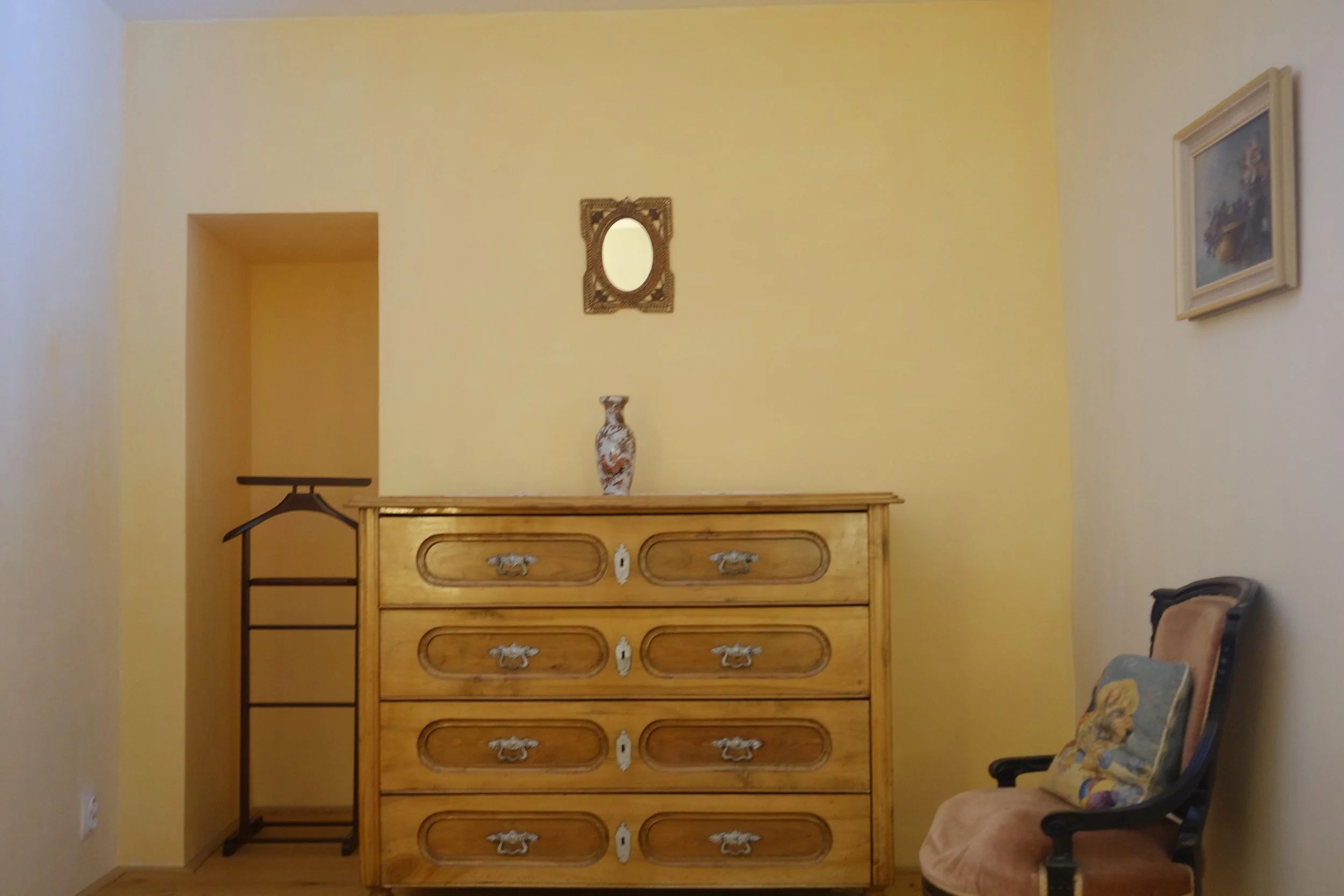 A wooden dresser with six drawers, topped with a vase, standing against a yellow wall. To the right, an ornate chair with a decorative pillow, and a small painting hanging on the wall.
