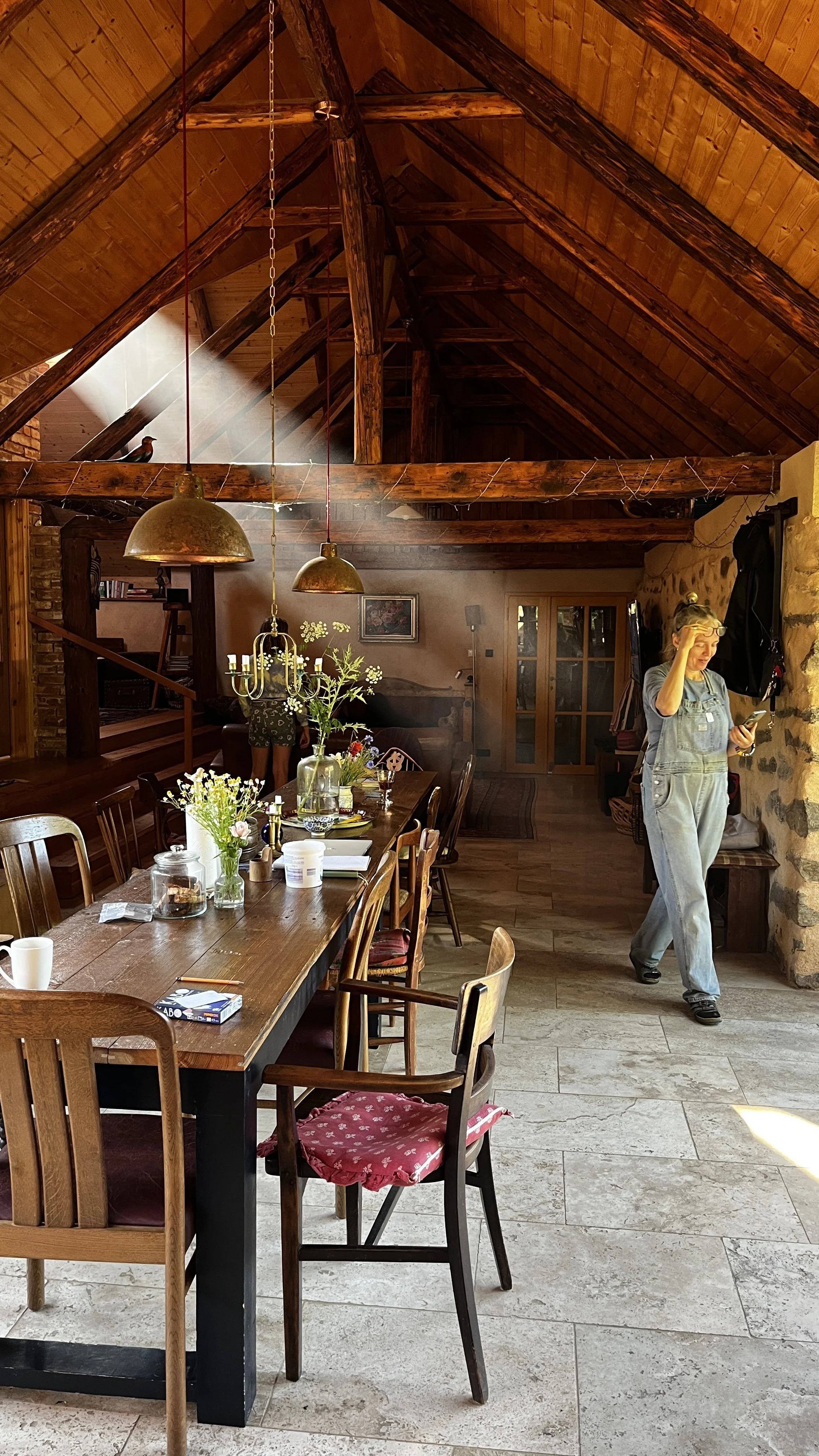 A cozy dining area with a long wooden table, surrounded by mixed wood chairs with some having red cushioned seats. The table is decorated with vases of flowers, candles, and various items. The room features wooden vaulted ceilings with exposed beams, stone walls, and sunlight streaming in. A woman in overalls is standing near the stone wall, looking at her phone, with a backpack hanging on the wall. There is a chandelier-style light fixture hanging above the table, and a beam with a bird perched on it is visible in the background.