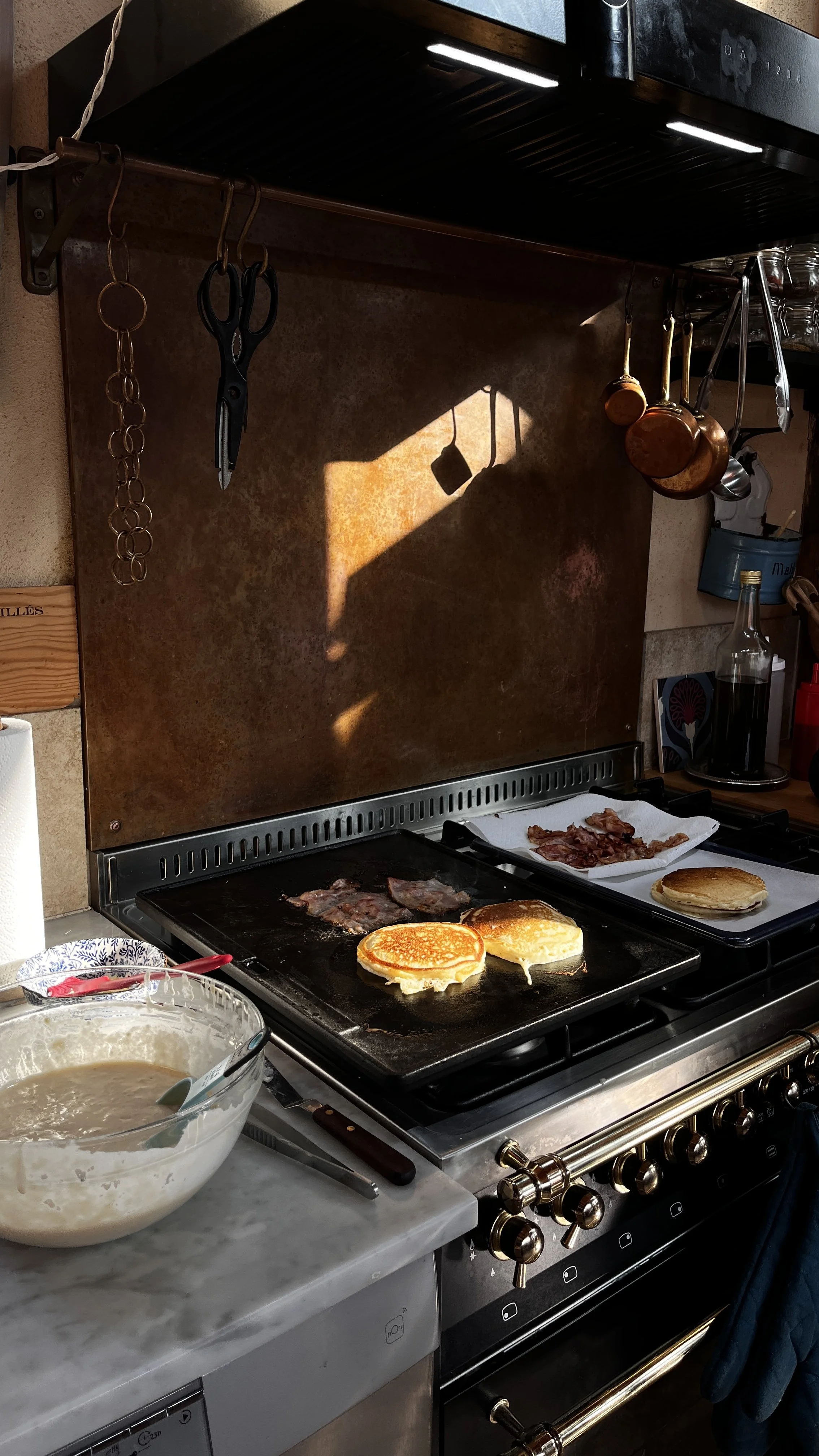 A stovetop with pancakes cooking, bacon on a paper towel, and a batter in a bowl nearby, with kitchen utensils hanging above.