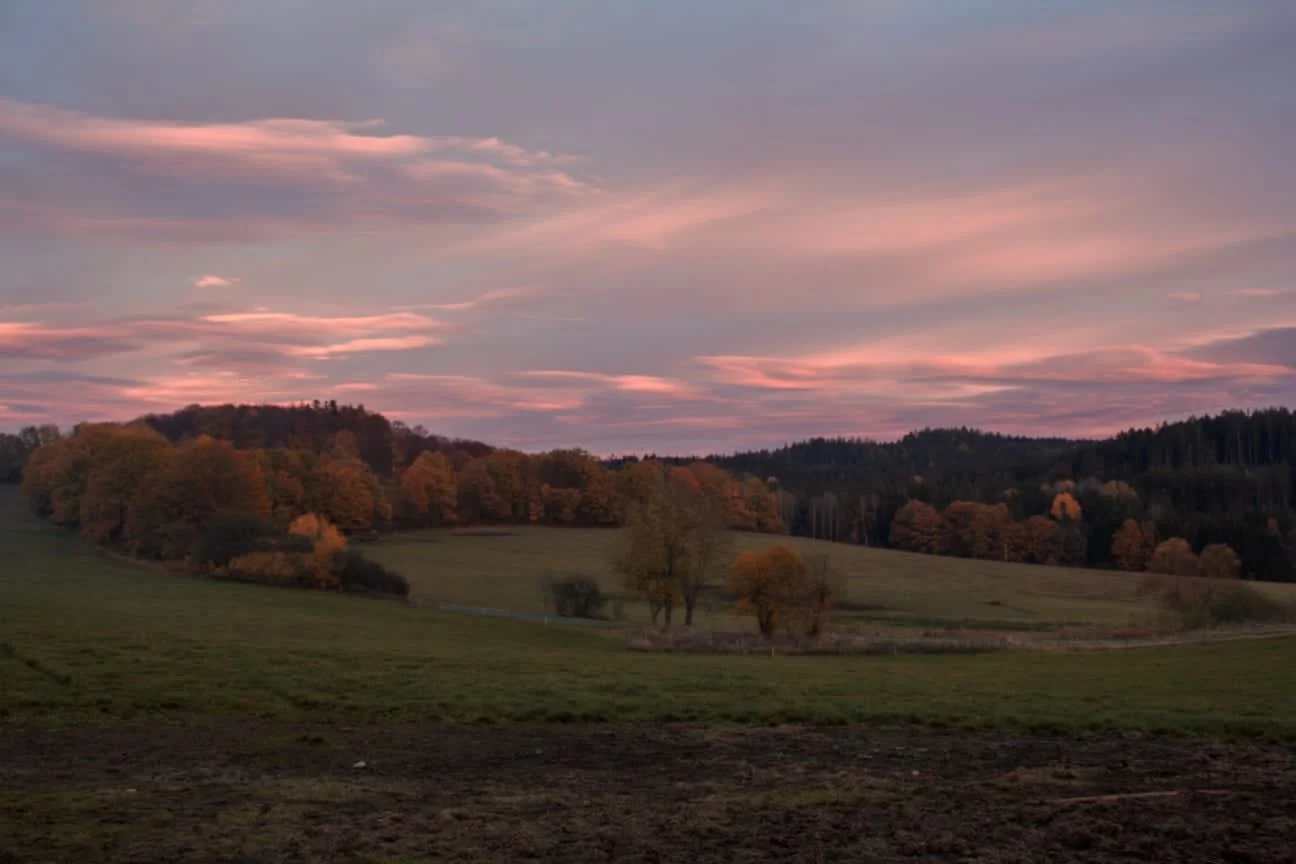 A scenic Czech countryside landscape during sunset, with rolling green hills, scattered trees in fall colors, and a pastel pink and purple sky with wispy clouds.