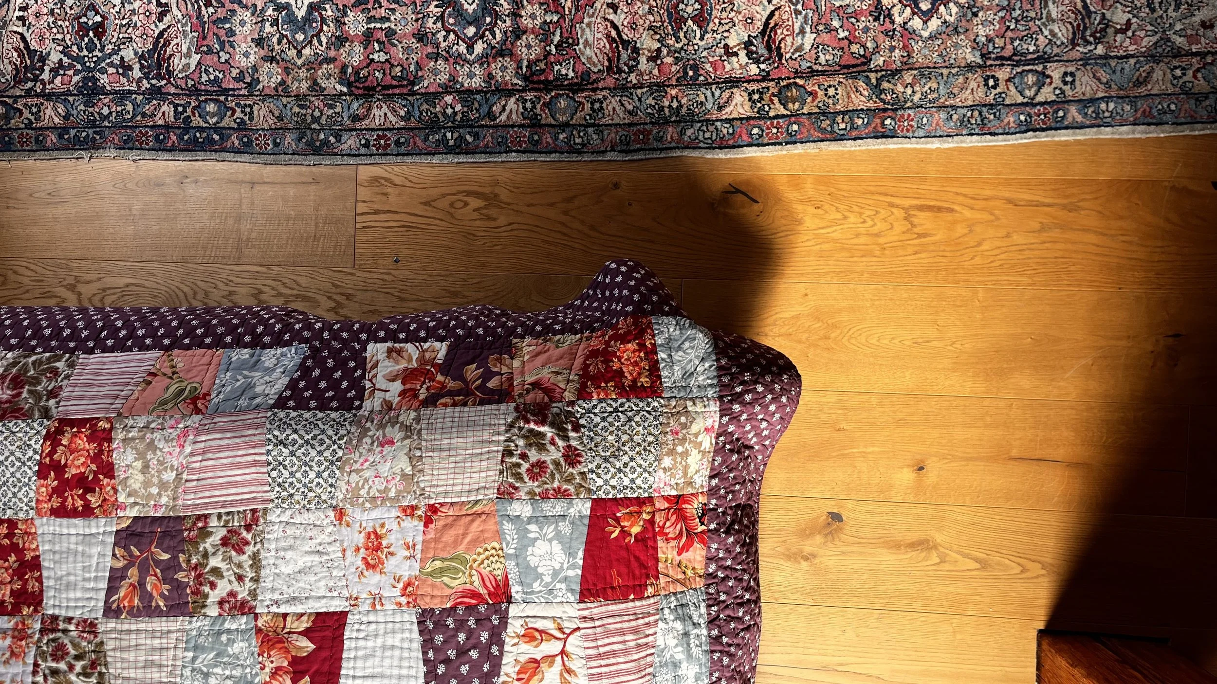 Overhead view of a patchwork quilt with floral patterns resting on a wooden floor, next to a colorful woven rug.