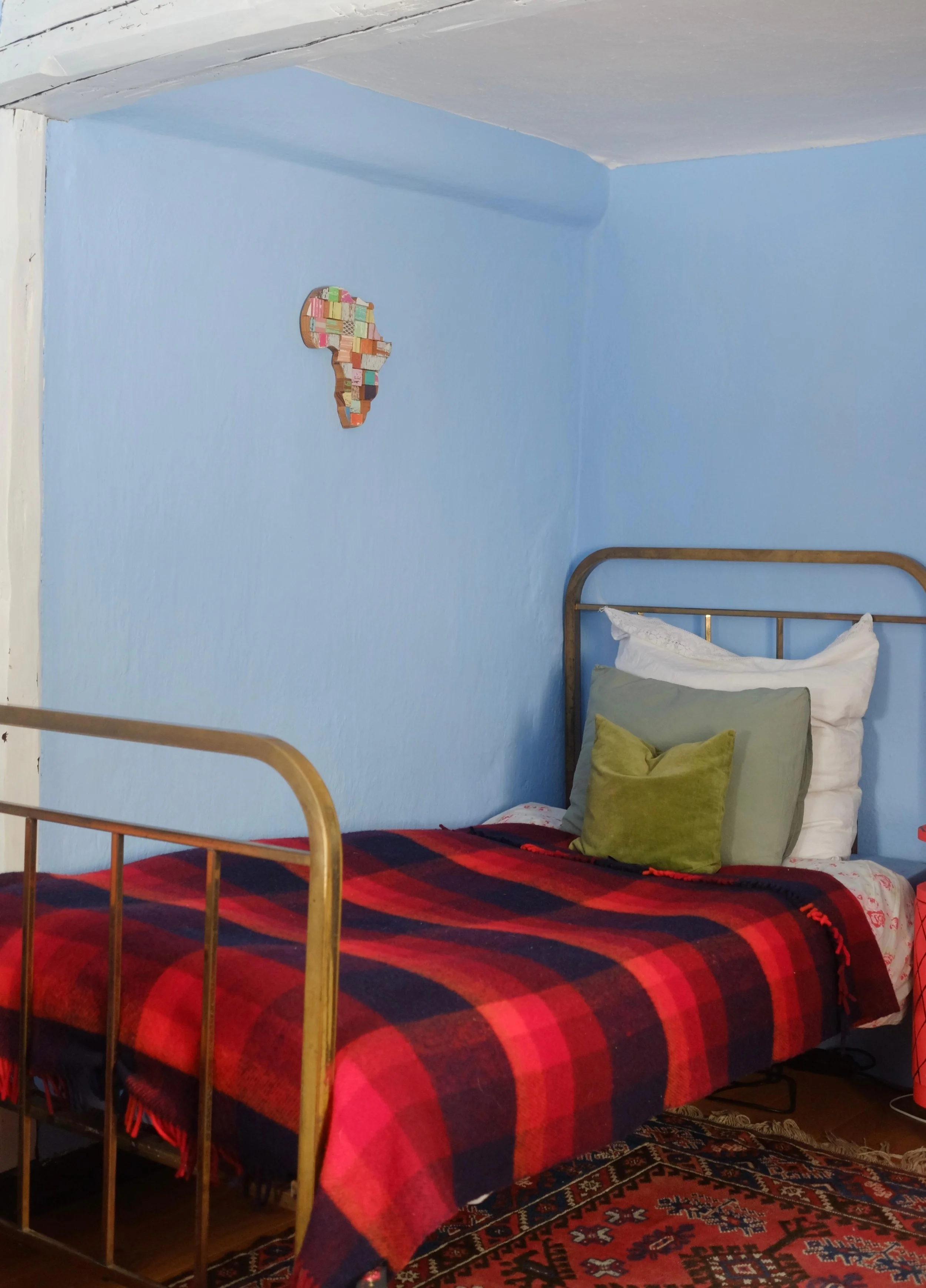 A bedroom with light blue walls, a brass bed with a red and black plaid blanket, pillows including white, gray, and green, and a colorful Africa-shaped wall decor made of small, different colored blocks.