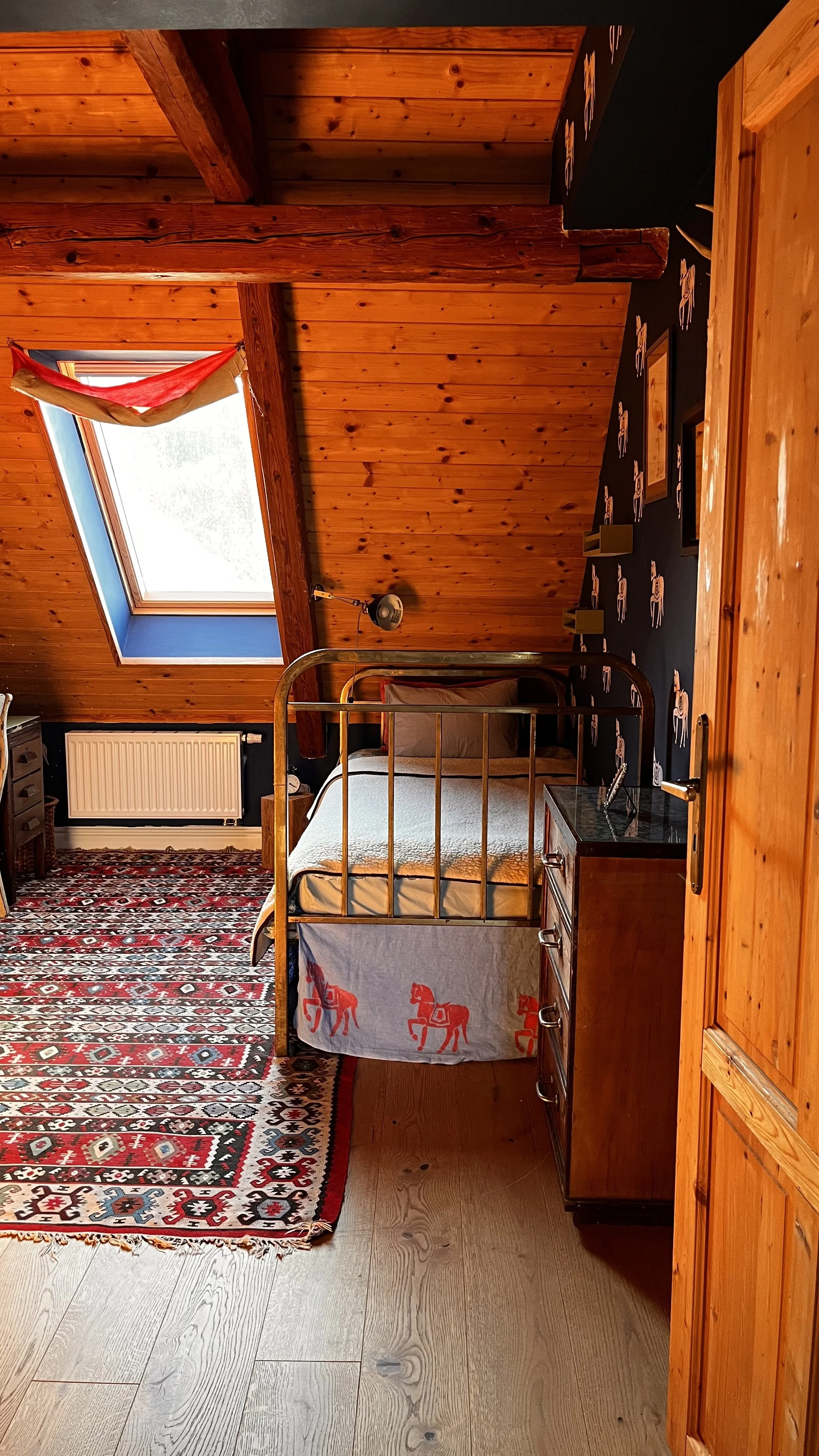 A cozy bedroom with wooden walls and ceiling, a skylight window with a red and yellow curtain, a patterned red, black, and white rug, a brass bed with a white bedspread featuring red horses, a black nightstand with a lamp, and a wooden wardrobe.