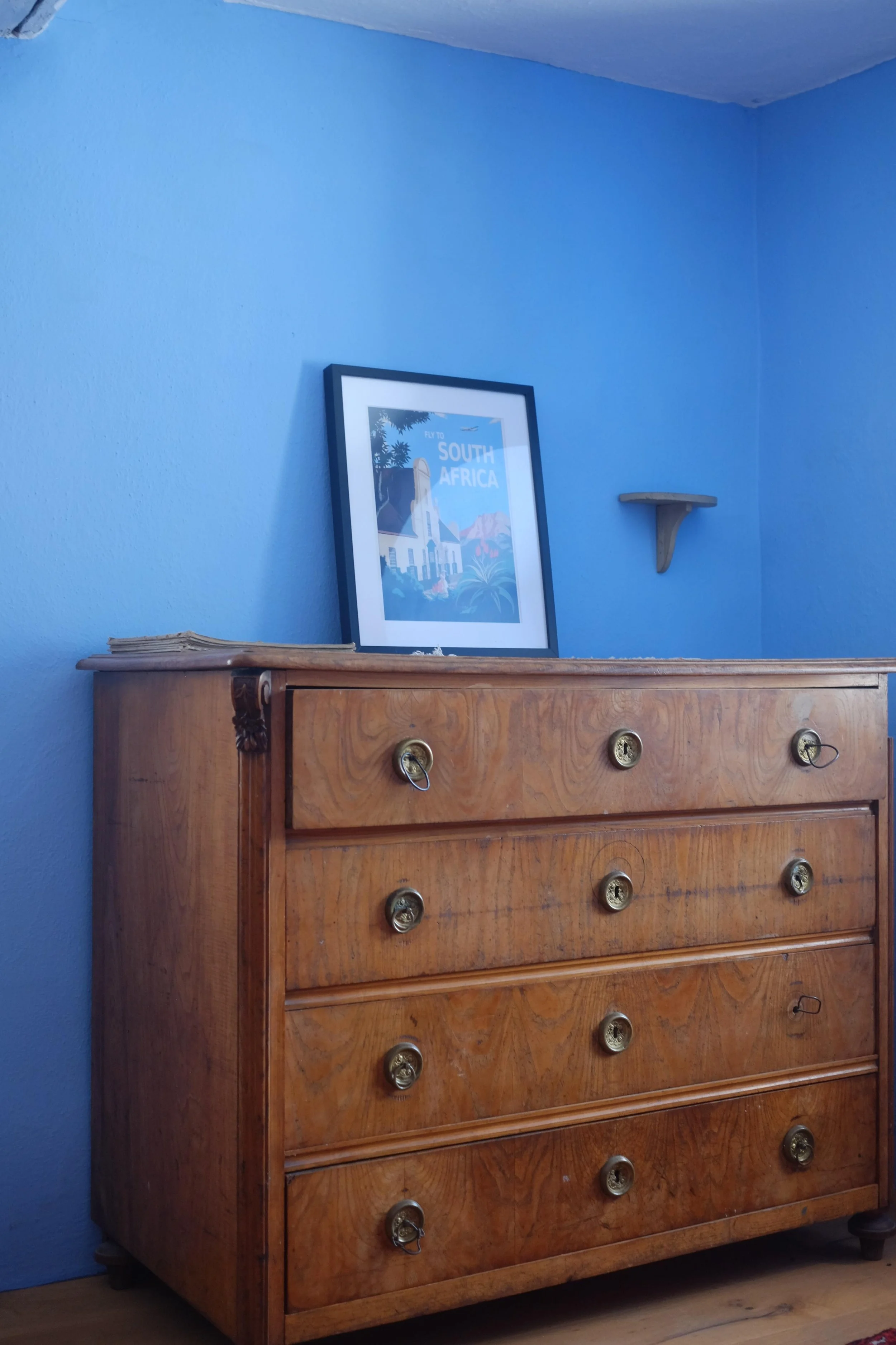 A wooden dresser with brass-colored round drawer handles positioned against a blue wall. A picture with travel illustration and the text 'To South Africa' rests atop the dresser. A small shelf is mounted on the wall.
