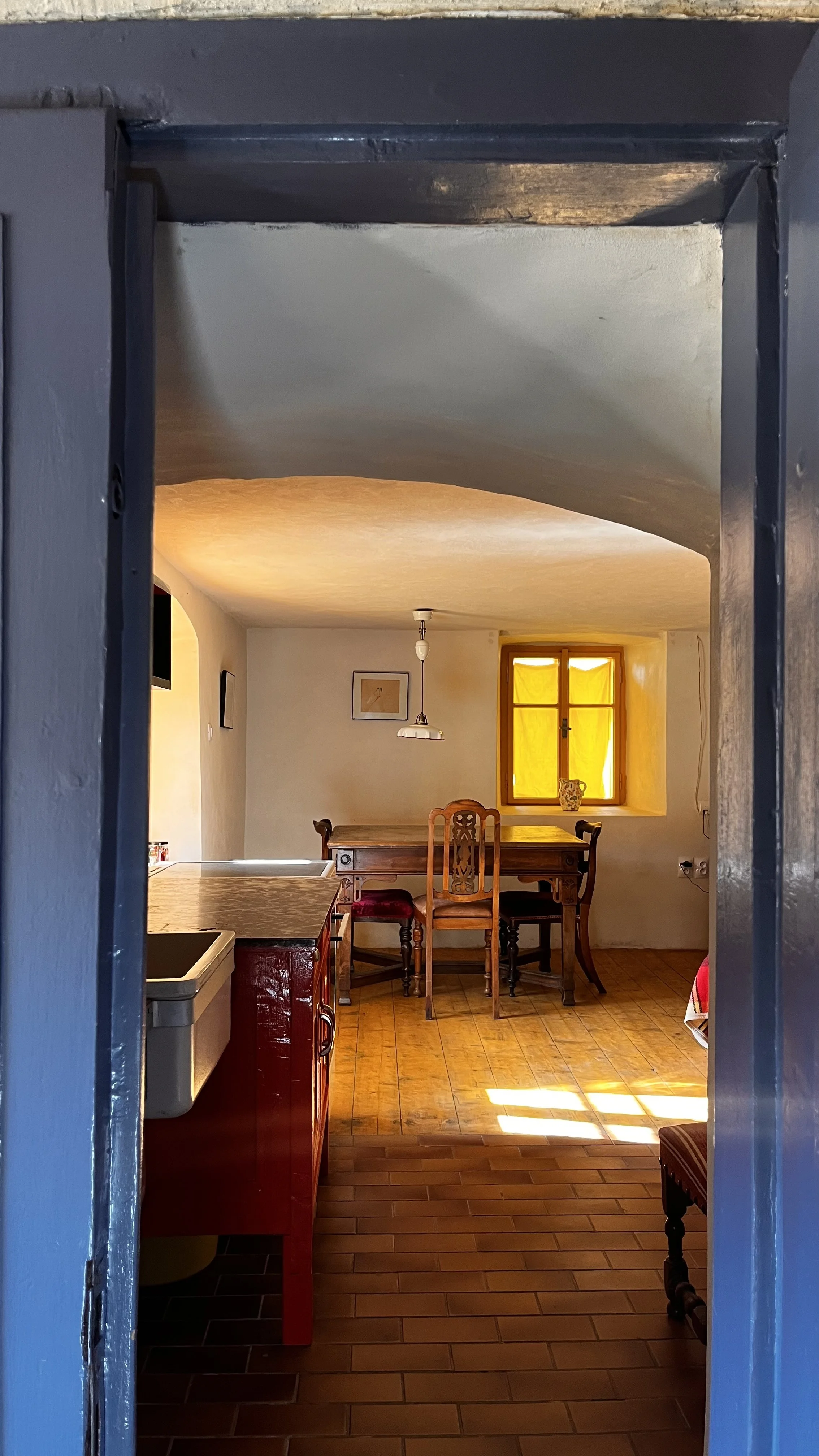 View through a small doorway into a cozy kitchen and dining area with wooden chairs, a yellow window, and warm lighting.