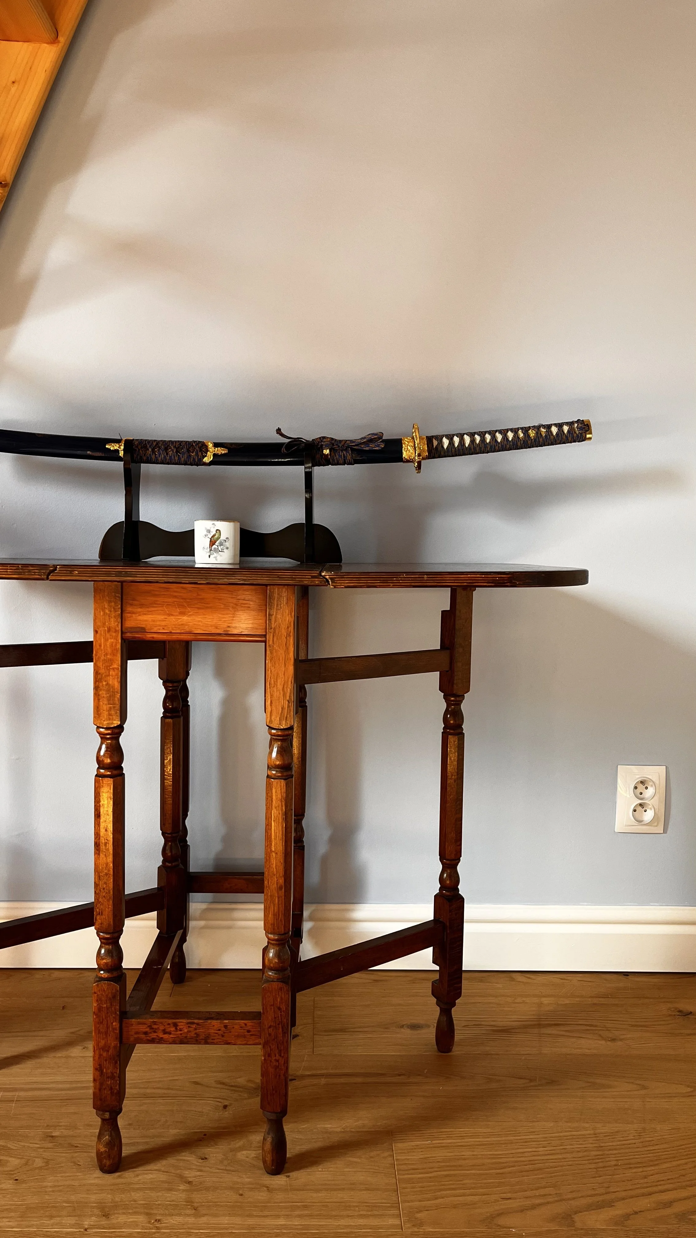 A katana sword displayed on a black stand on a wooden table against a light gray wall.