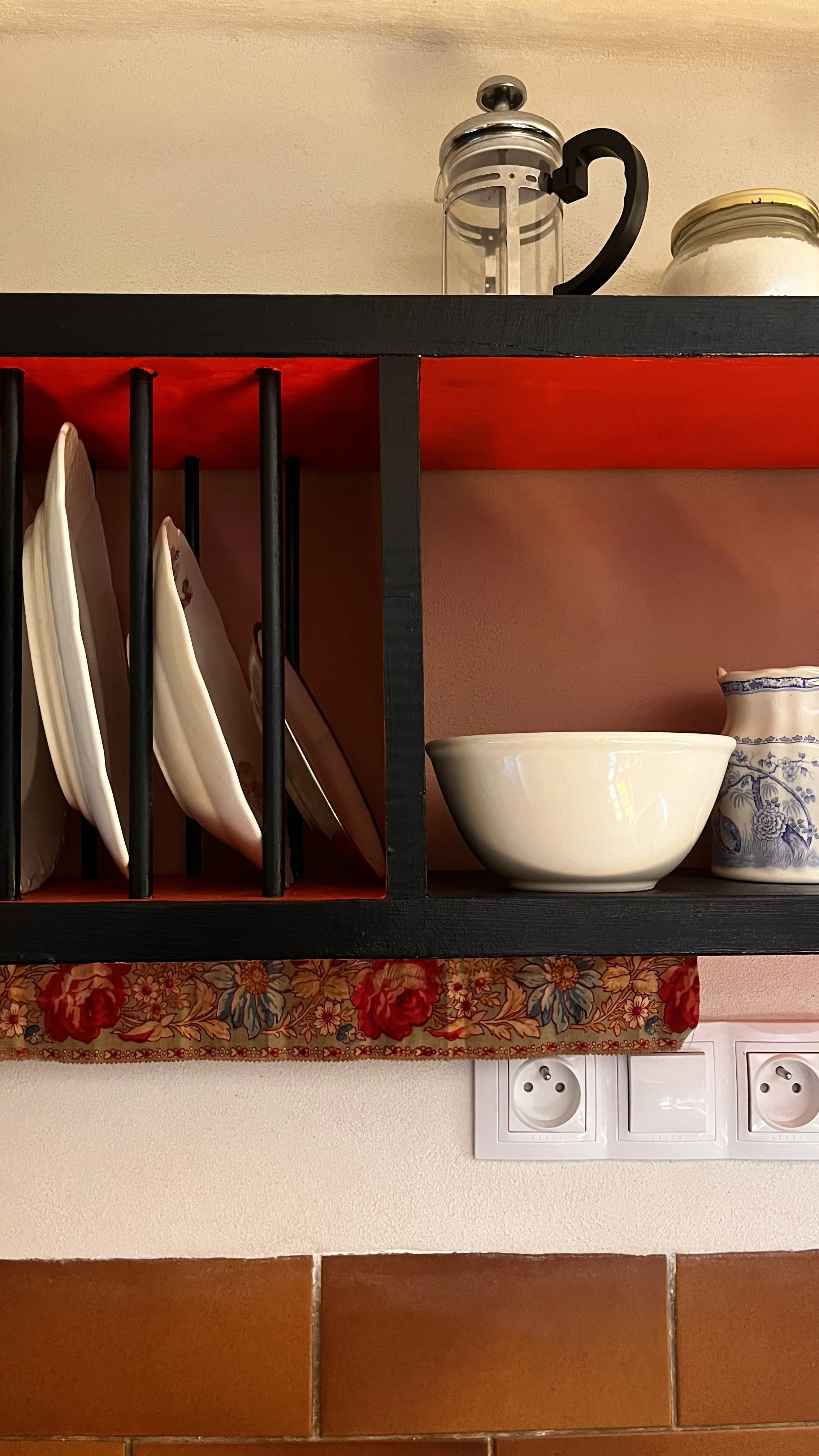 Close-up of a kitchen shelf with plates, bowls, and decorative jars, with a floral curtain below the shelf and electrical outlets on the wall.