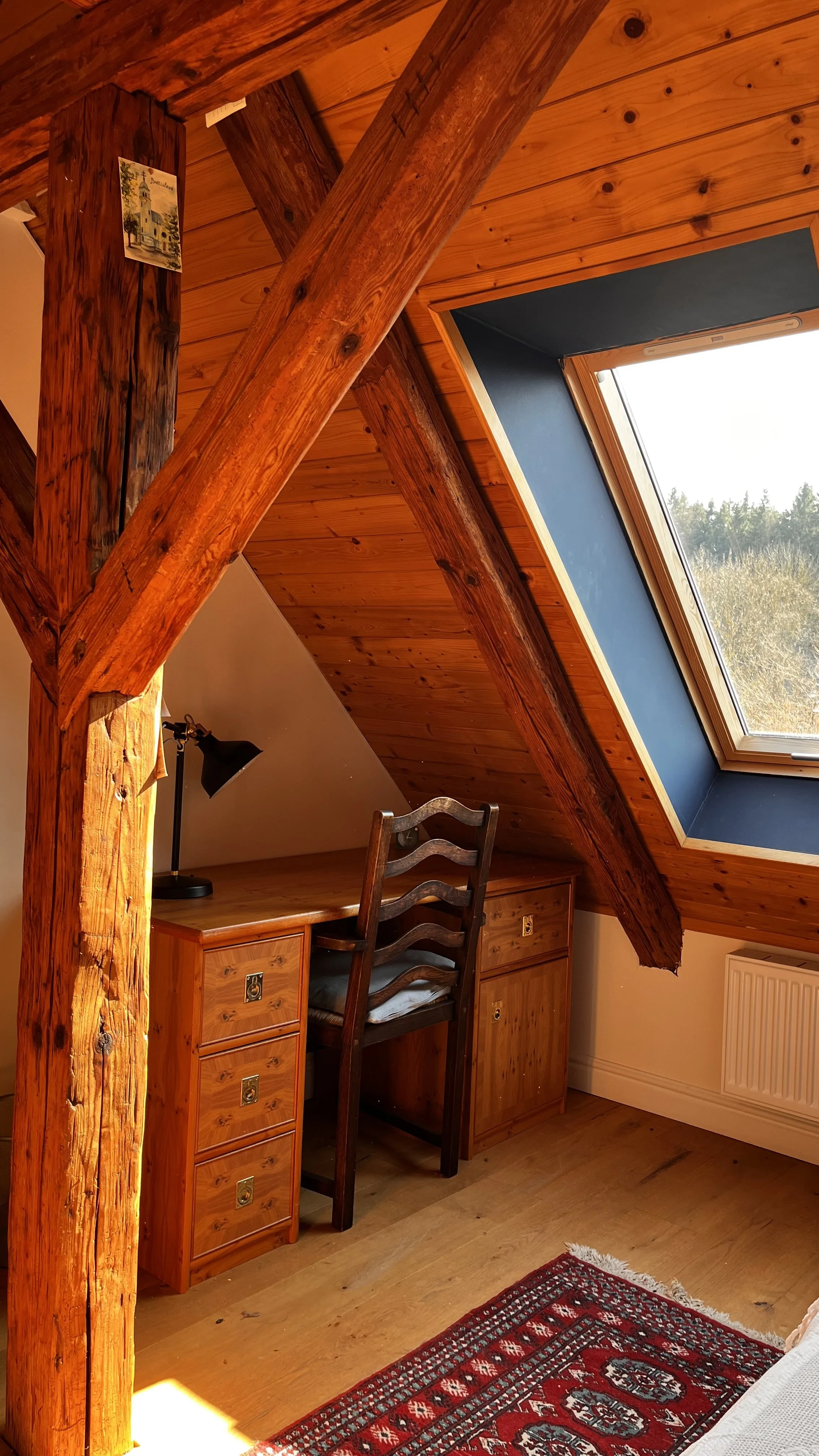 A cozy attic room with wooden beams, a sloped wooden ceiling with a large skylight window, a wooden desk with a desk lamp, a wooden chair, and a red patterned rug.