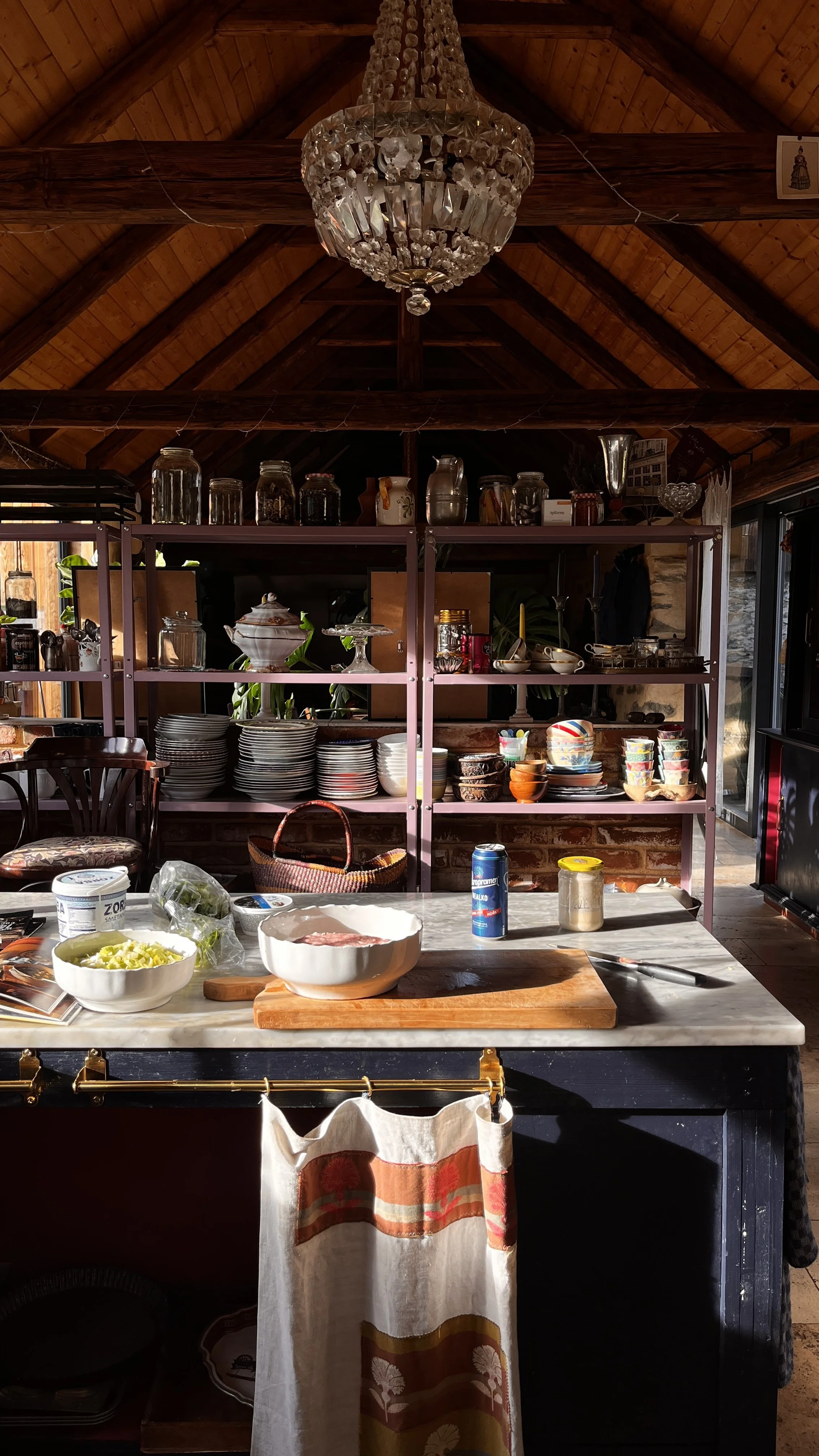 Kitchen with a marble countertop, bowls of chopped vegetables, a cutting board with a piece of meat, and metal kitchen utensils. Behind, open shelves with dishes, bowls, and jars, and a chandelier hanging from a wooden vaulted ceiling.