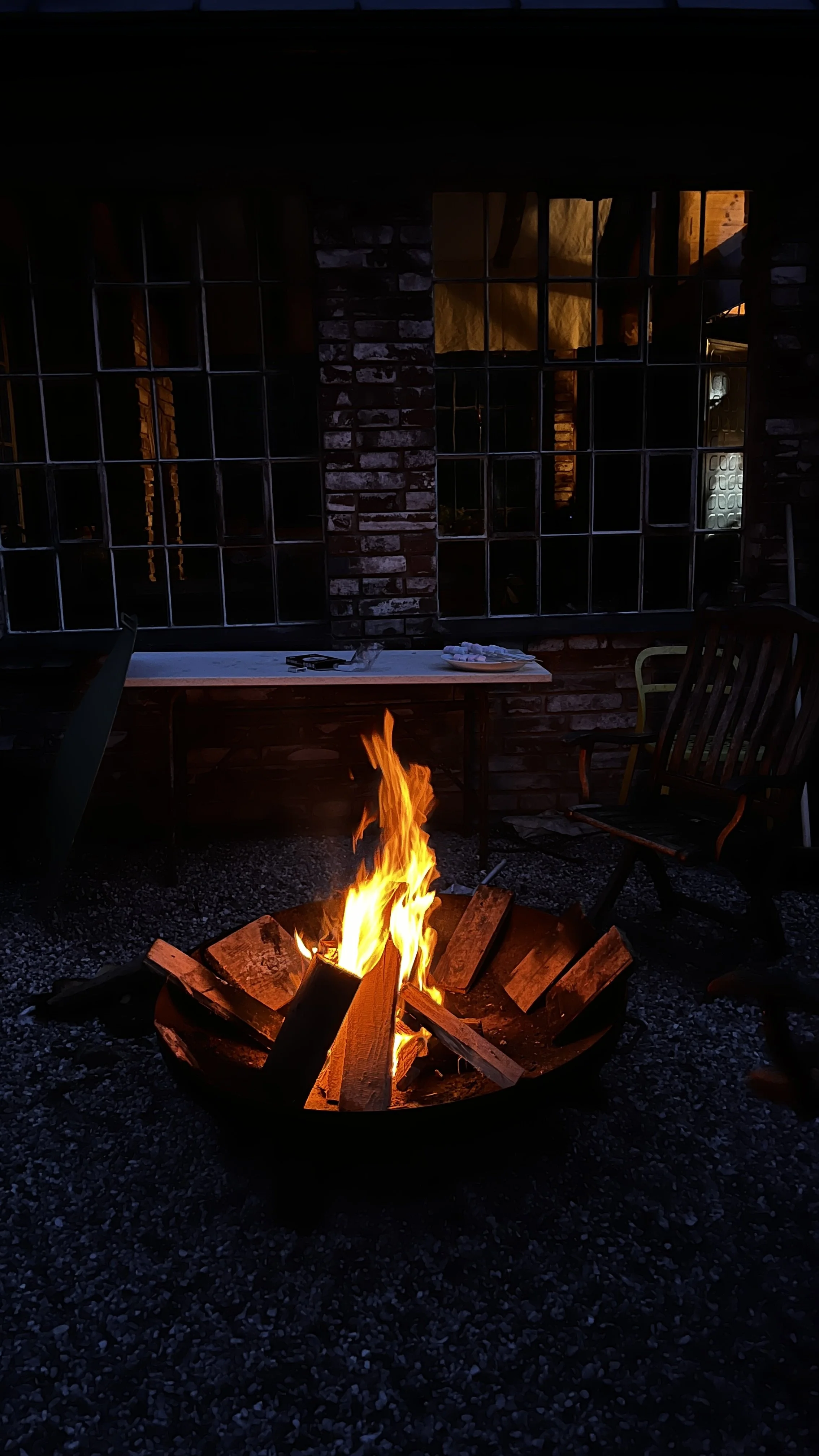 A fire burning inside a fire pit outside a building at night, with chairs and a table in the background.