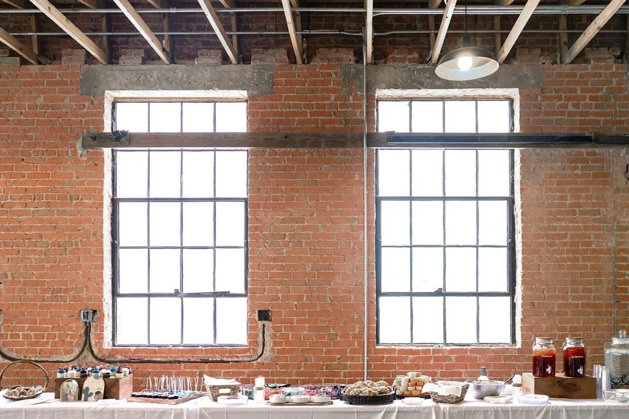 Indoor party setup with a table of snacks and drinks against a brick wall with two large windows.