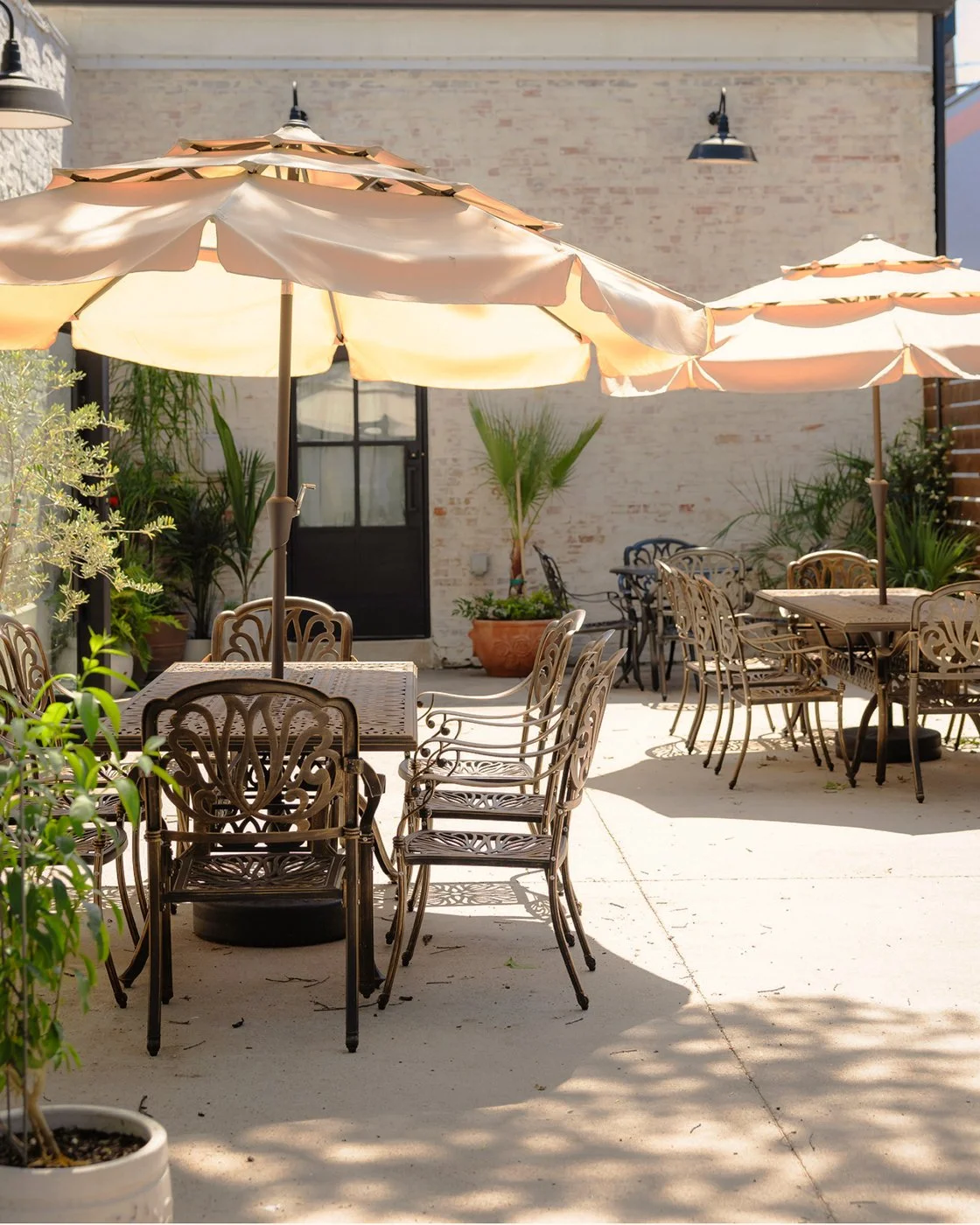 Outdoor patio with metal tables and chairs, large beige umbrellas, potted plants, and brick wall in the background.