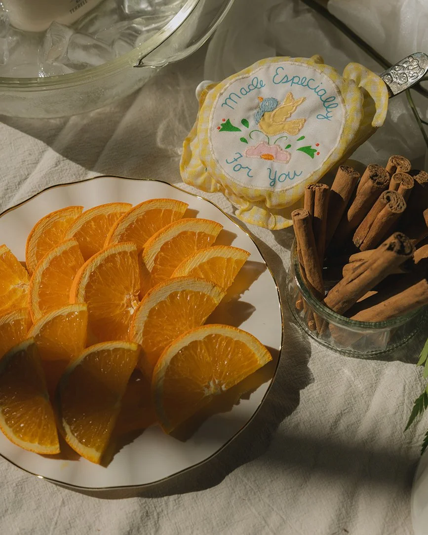 A white plate with orange slices arranged in three rows. A glass jar containing cinnamon sticks is on the right side. Part of a fabric with a yellow and white checkered cloth and a cloth covering embroidered with a unicorn and the words "Made Especially for You" is visible. A glass container with ice and a metal spoon is in the background.
