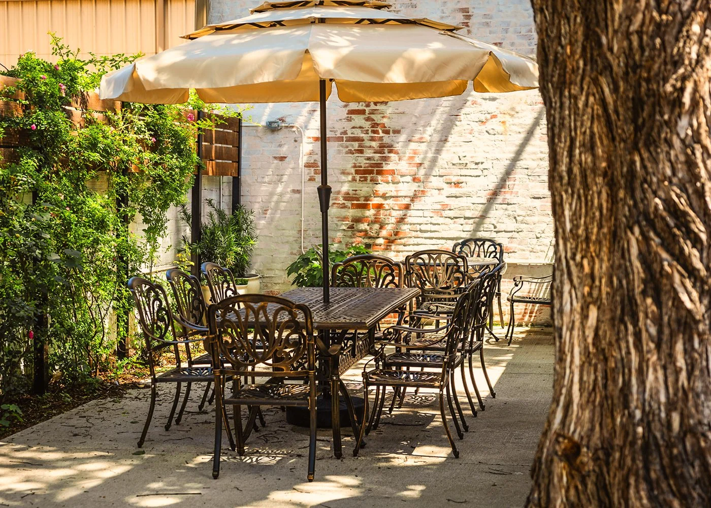 Outdoor patio with a large dining table surrounded by metal chairs, shaded by a beige and brown patio umbrella, with a tree in the foreground, brick wall and greenery along the side.