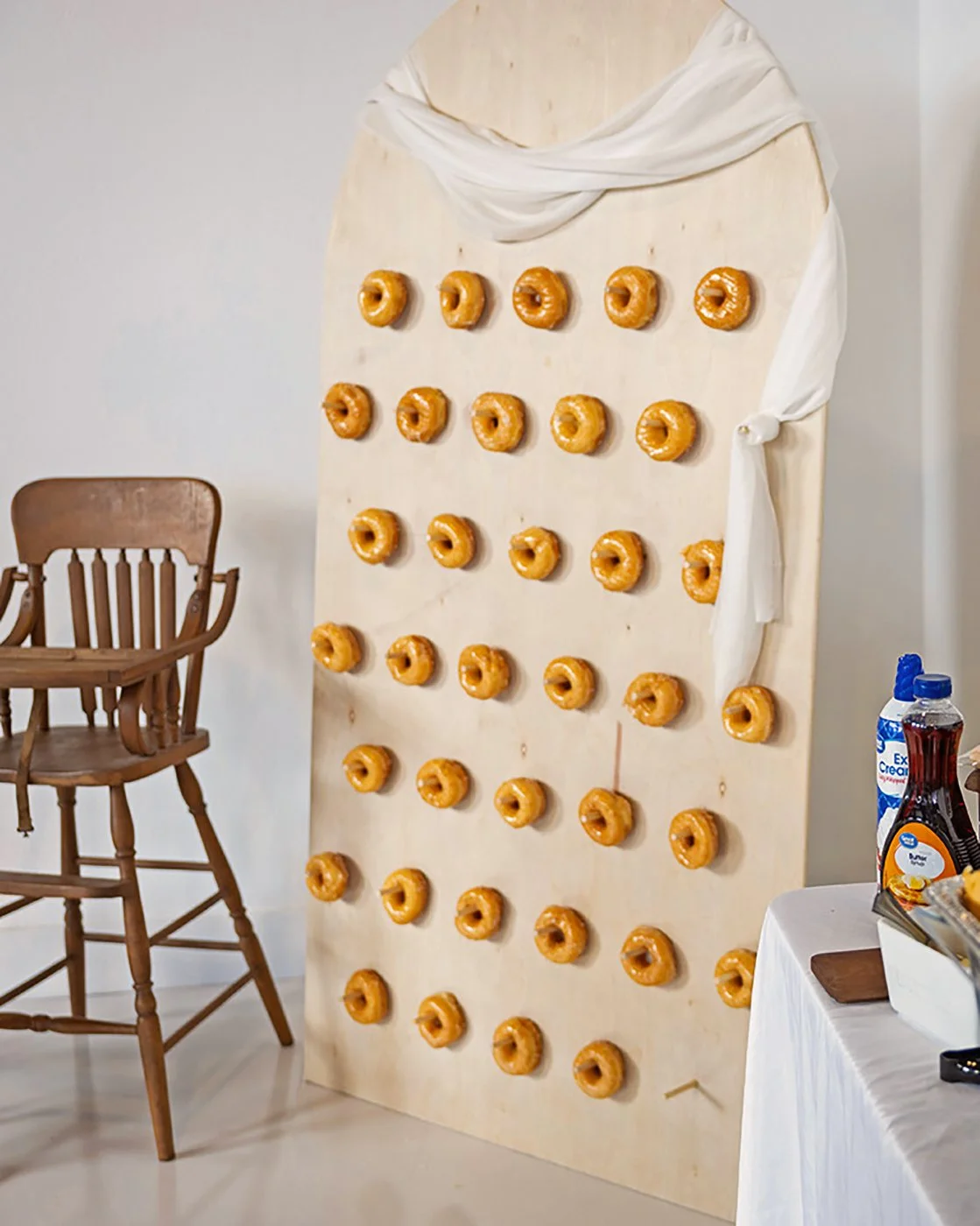 A tall wooden board with multiple rows of glazed donuts attached. The board is draped with white cloth and is situated next to a wooden chair and a table with various items.