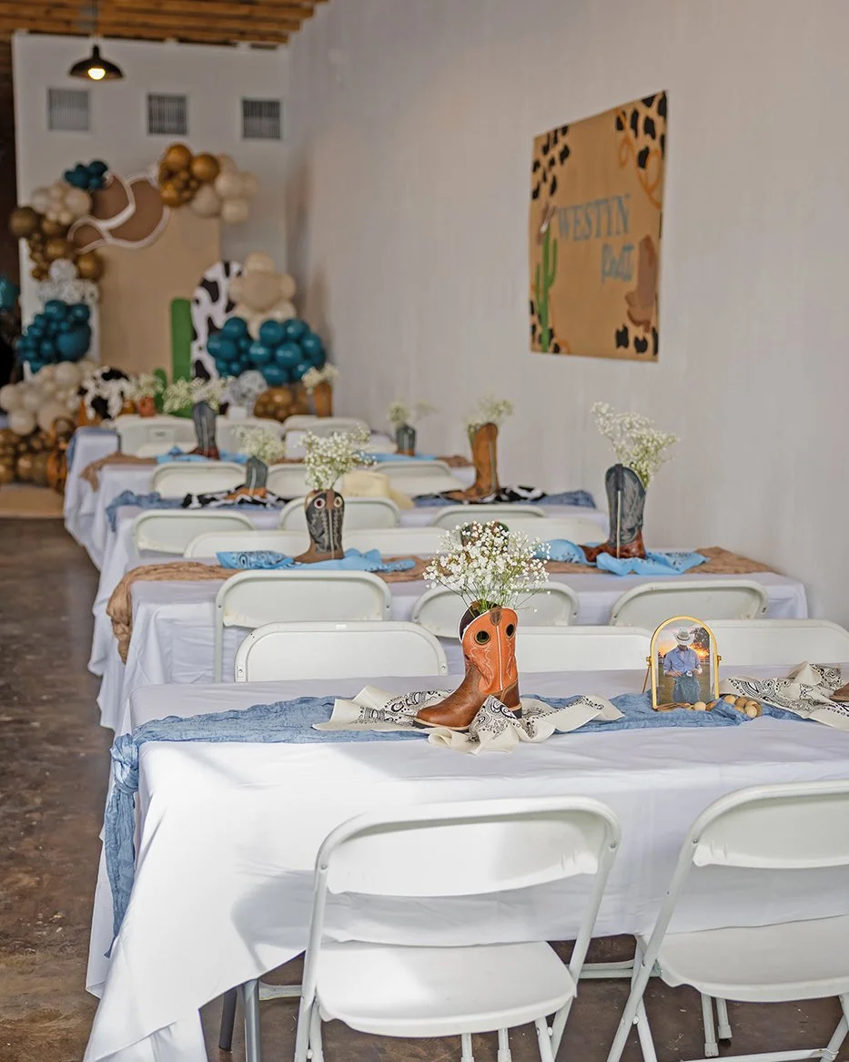 Long dining tables decorated with cowboy boot centerpieces and white flowers, set for a Western-themed party or event, with a balloon backdrop and Western decor in the background.