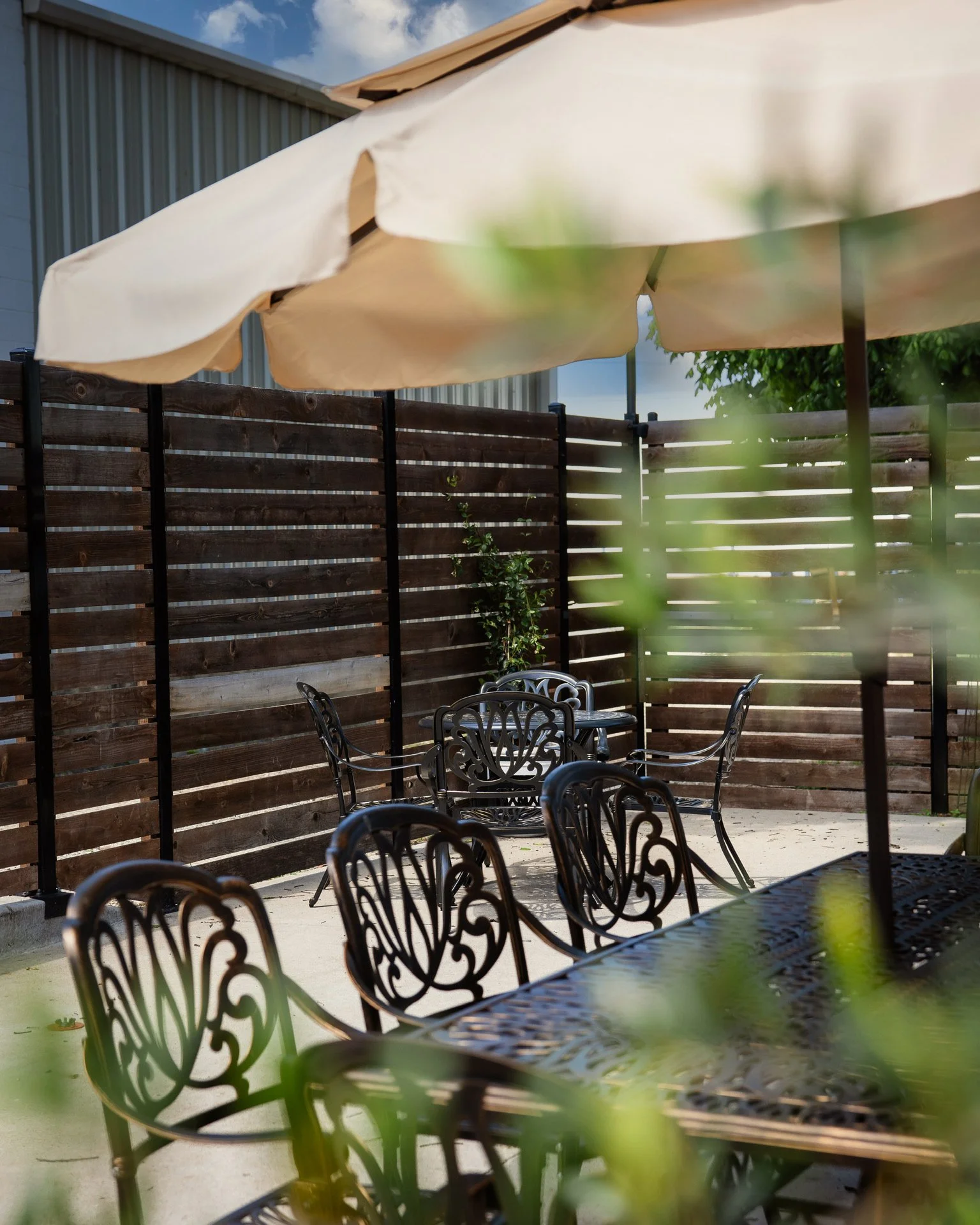 Outdoor patio with black metal chairs around a table, a large cream-colored umbrella, and a wooden privacy fence, with a building partly visible and blue sky with clouds above.