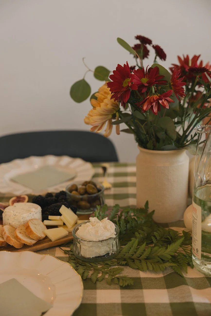 A table set with a green and white checkered tablecloth, holding a bouquet of red, yellow, and orange flowers in a beige ceramic vase, a plate with cheese and crackers, a small bowl of green olives, a jar with a creamy spread, and a large bottle of water. A black chair is visible in the background.