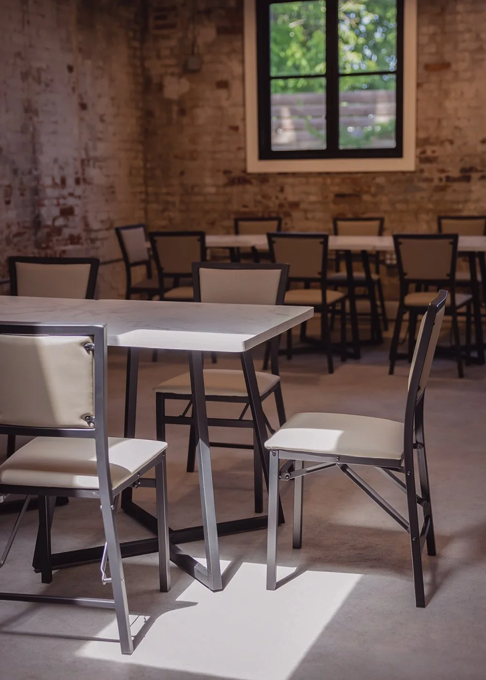 Empty dining area with modern chairs and tables in a room with brick walls and a window showing greenery outside.
