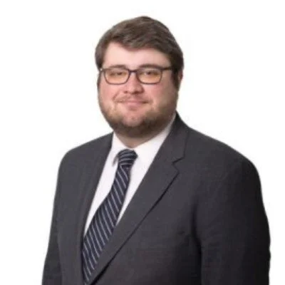 Professional man with glasses, dark hair, and beard wearing a dark suit, white shirt, and striped tie, smiling against a white background.