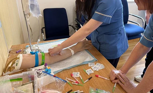 Medical training simulation with a mannequin arm on a table and medical professionals practicing injections
