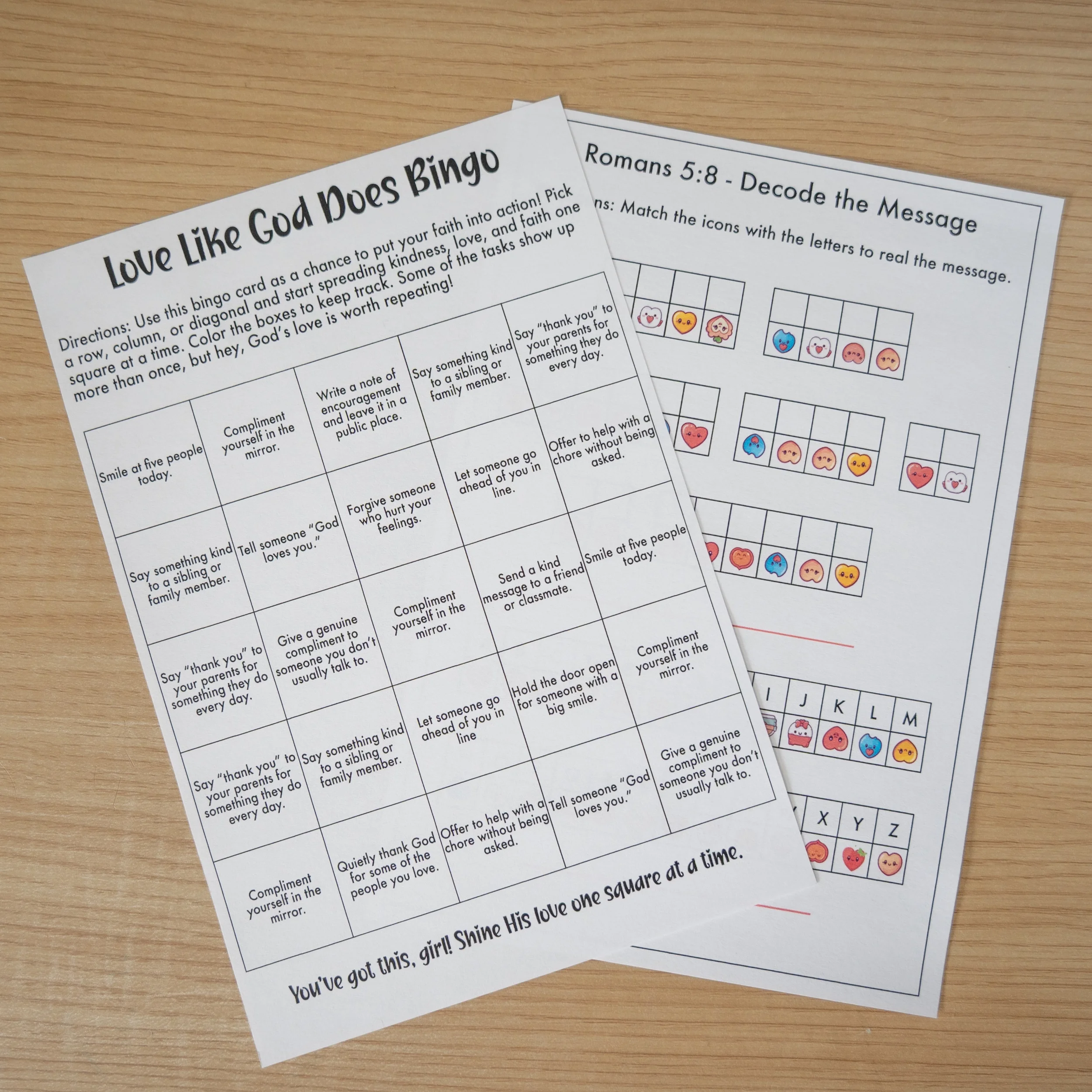 Two printed sheets of Bible-themed activities and games on a wooden surface; one sheet titled 'Love Like God Does Bingo' with instructions and a grid of tasks, the other titled 'Romans 5:8 - Decode the Message' with puzzles and matching icons.