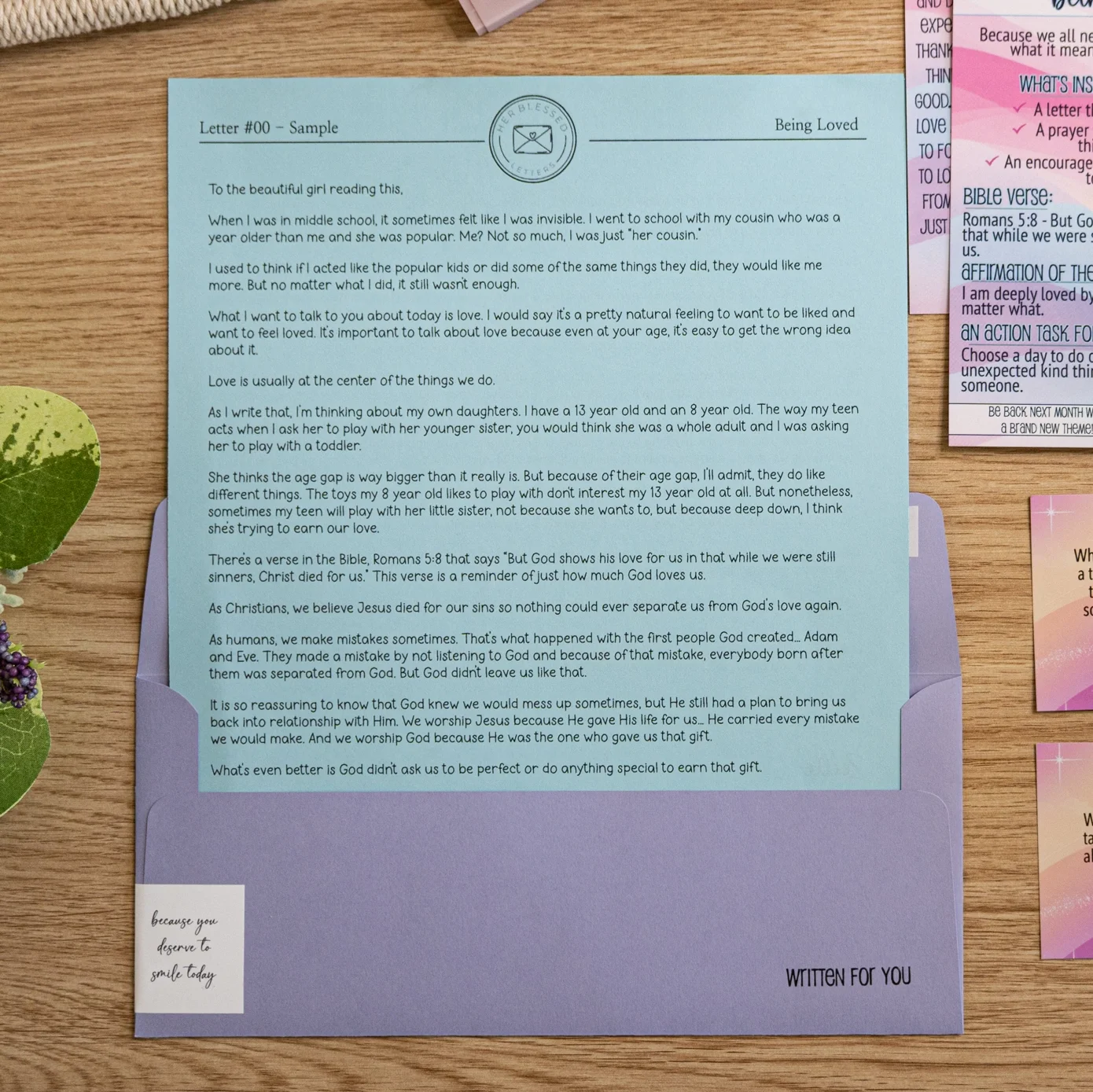 A blue letter on top of an envelope with a purple flap, situated on a wooden table. The letter contains a message about love, faith, and forgiveness, referencing the Bible verse Romans 5:8, and discusses relationships and God's love. A small note sticker on the envelope reads, "because you deserve to smile today."