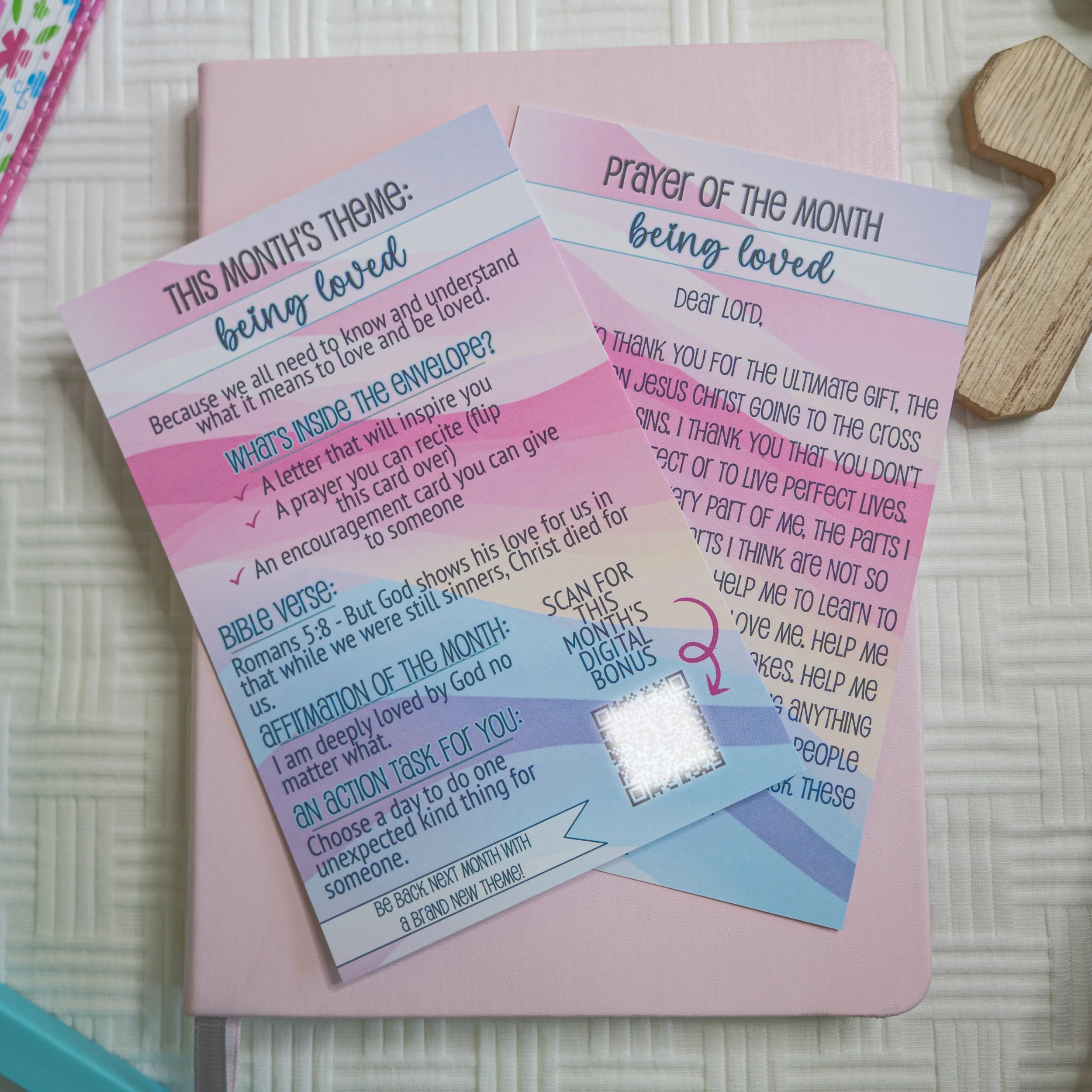 Two printed prayer cards titled 'Being Loved' and 'Prayer of the Month,' placed on a pink closed journal, with a wooden cross and other stationery nearby on a white textured surface.