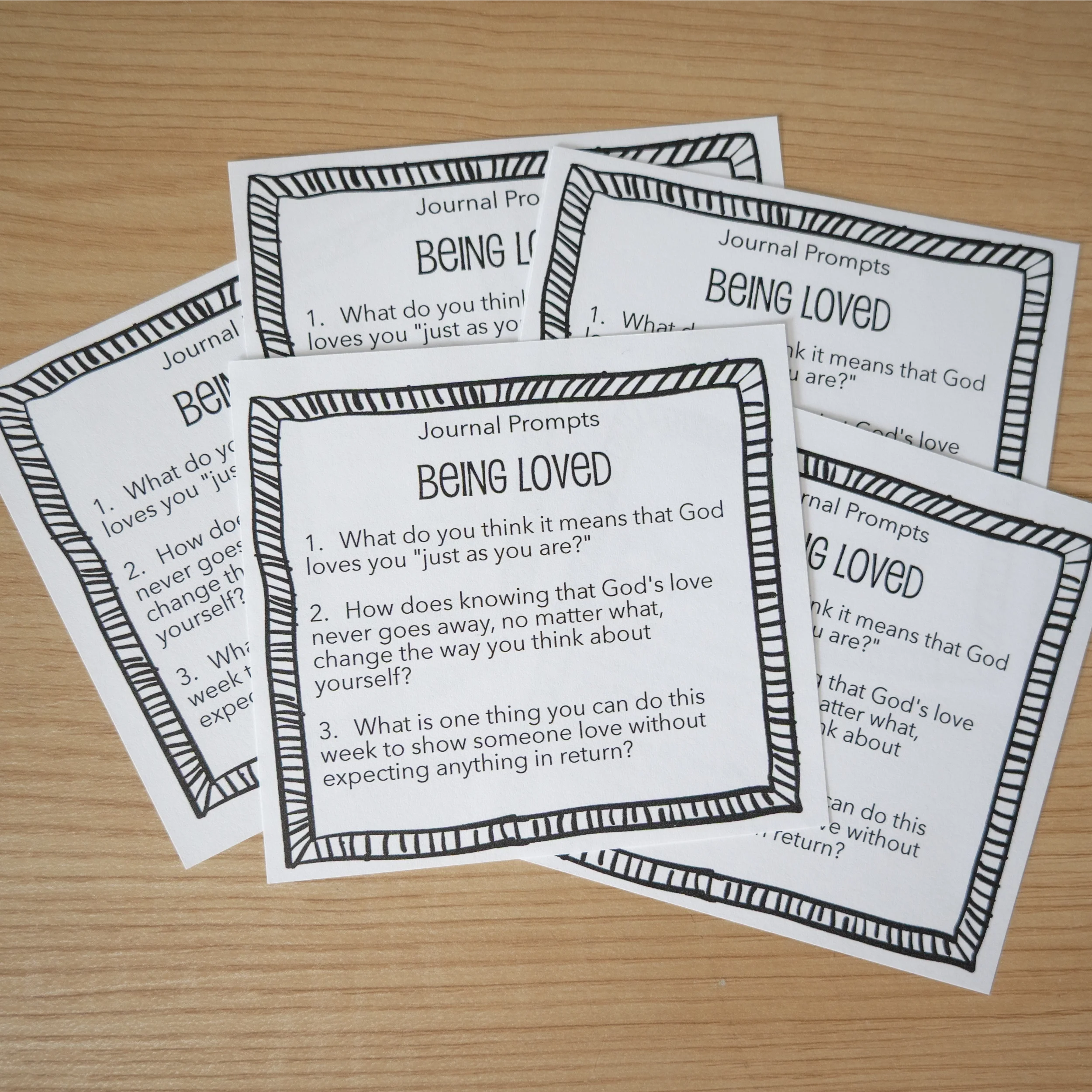 Several printed sheets of paper with journal prompts titled 'Being Loved' placed on a wooden surface.