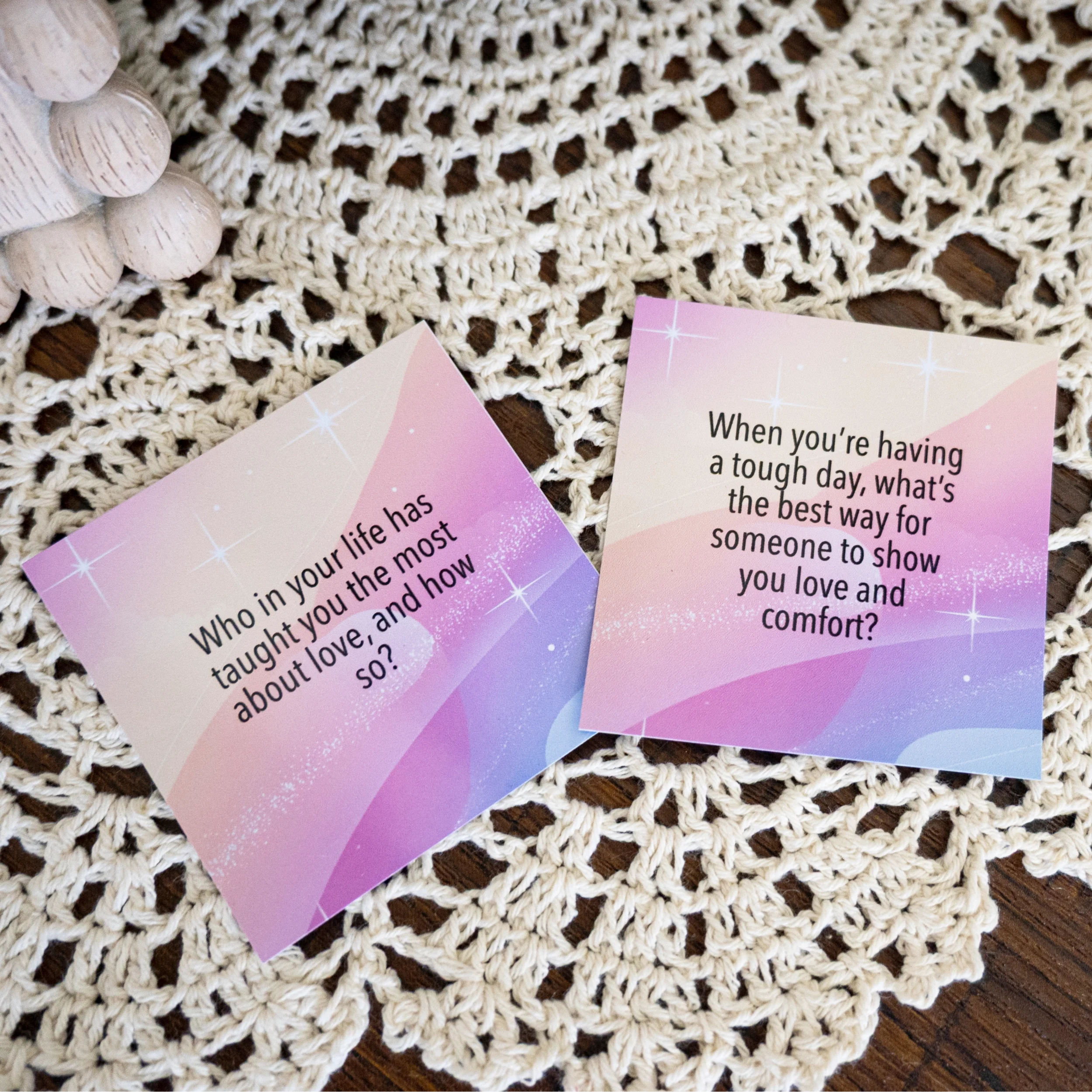 Two colorful cards with inspirational questions placed on a crocheted white tablecloth, with a decorative white beaded object partially visible on the top left.