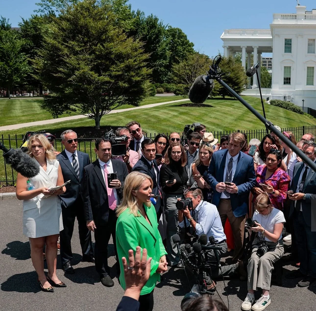 A woman in a bright green blazer speaking to a group of journalists outside the White House, equipped with cameras and microphones, under a clear sky with trees and the White House in the background.