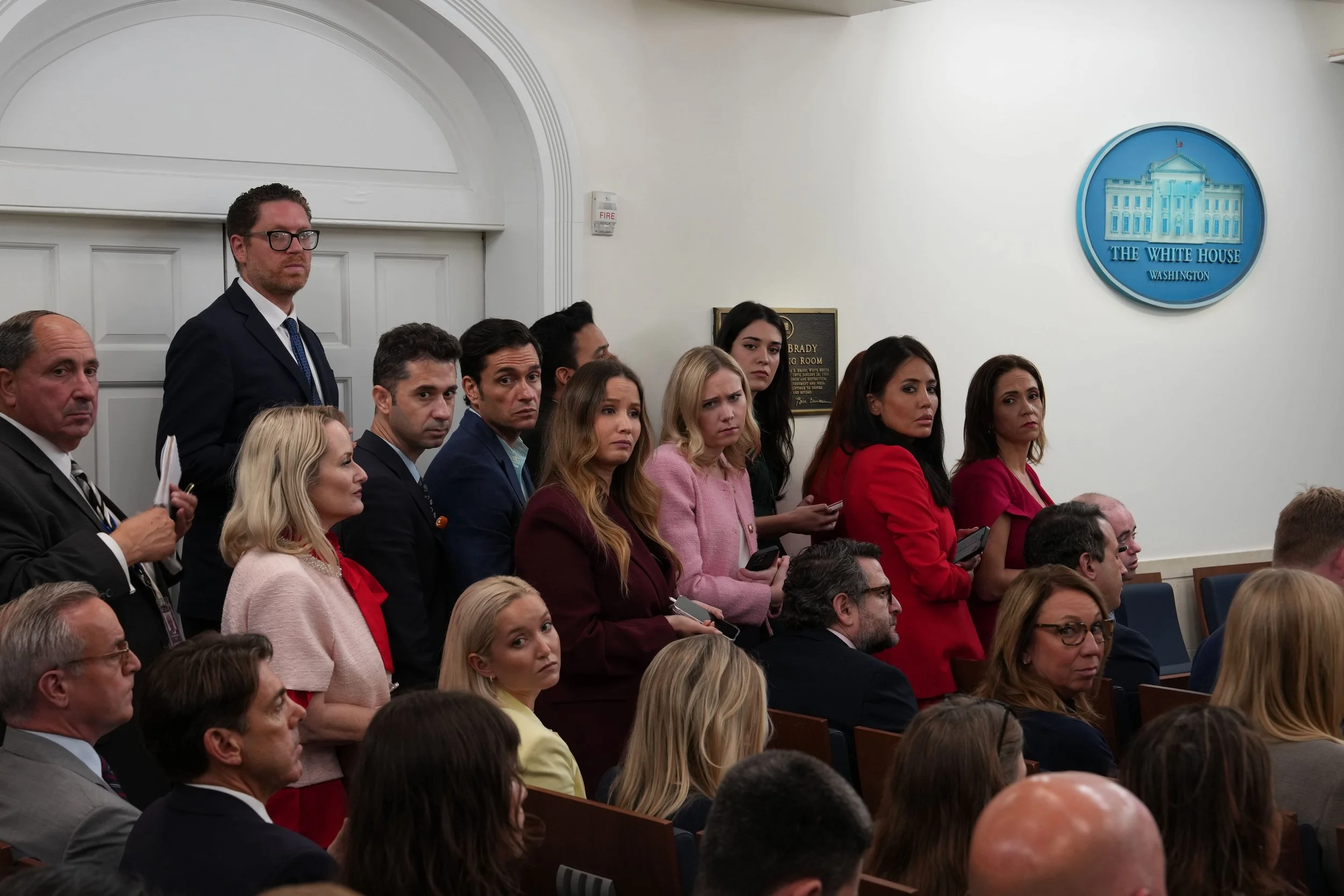 Group of people standing and sitting in a room with a United States White House emblem on the wall, indicating a formal government or press event.