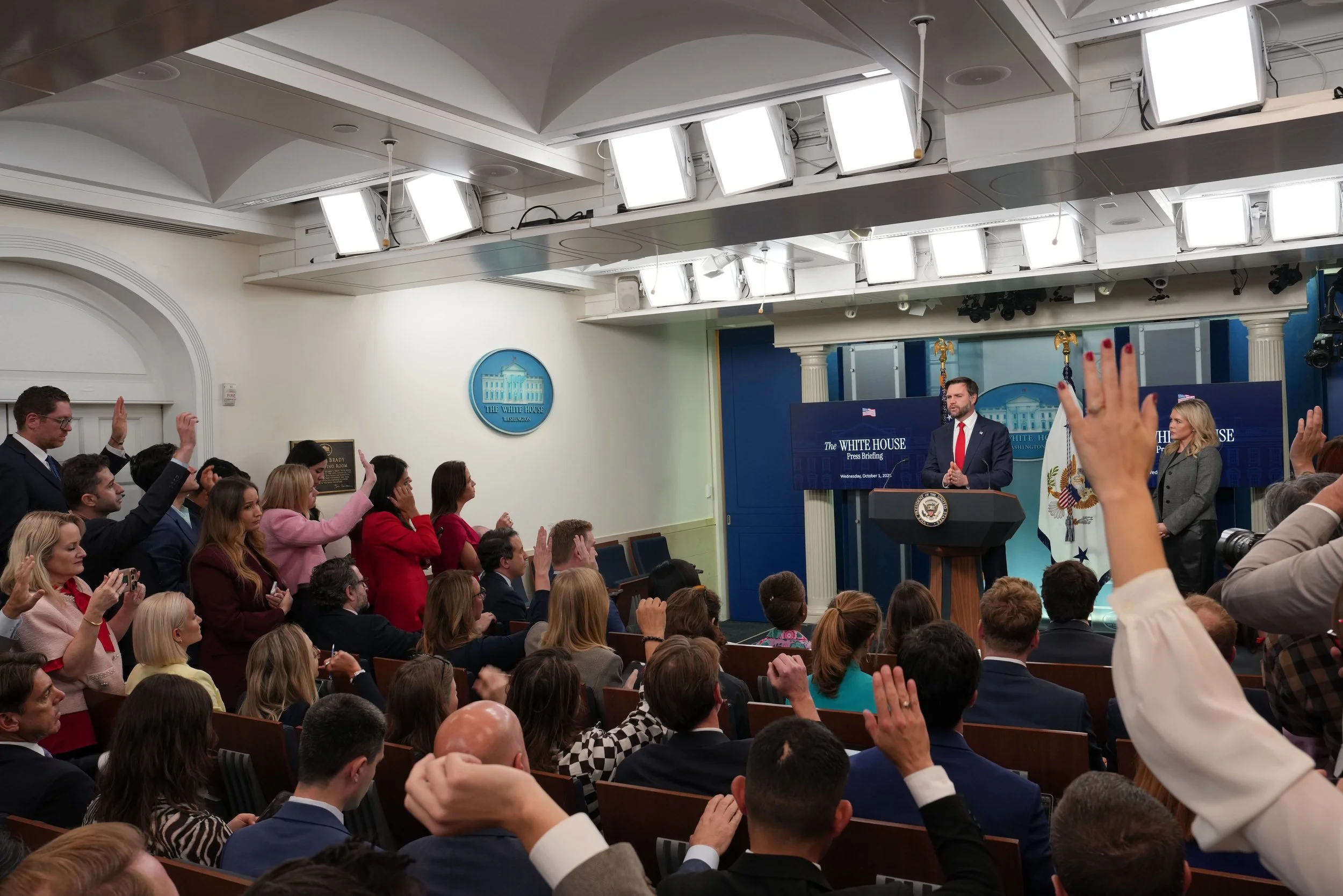 Press briefing at the White House with an audience of diverse individuals, some raising their hands, and a speaker at a podium with the White House emblem, U.S. flag, and a large screen behind him.