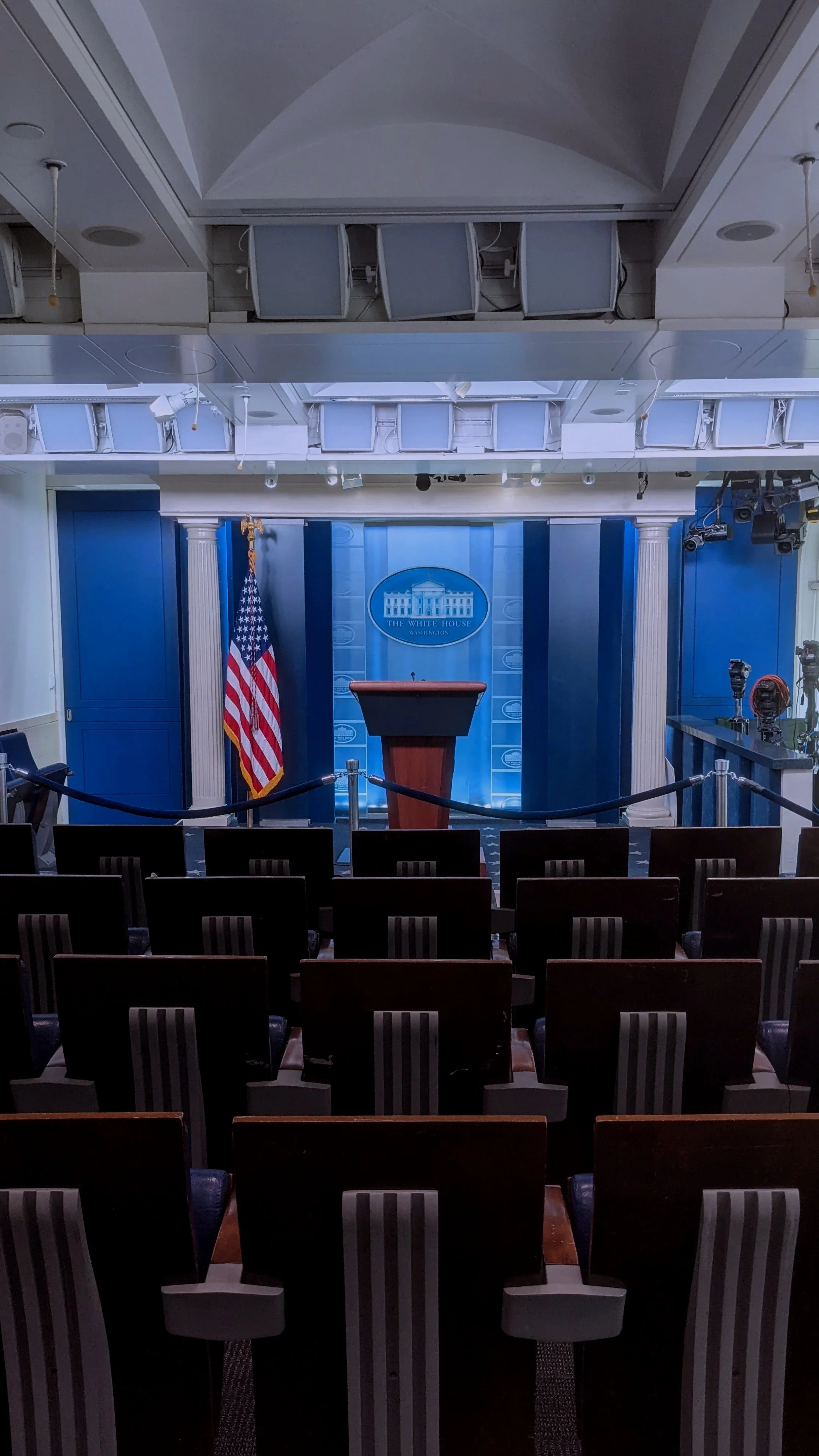 Empty press briefing room at the White House with podium, American flag, chairs, and camera equipment.