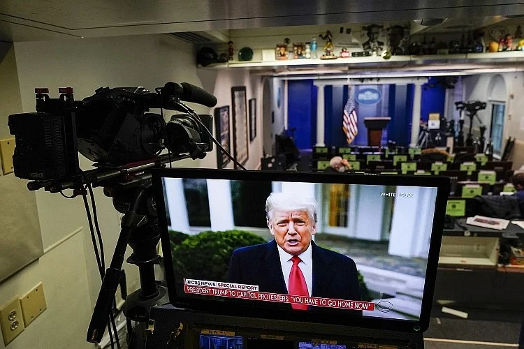 A television screen showing a news report of former President Donald Trump speaking outside the White House, with a crowd of protesters, in a room set up for a press conference with rows of desks and people working at computers.