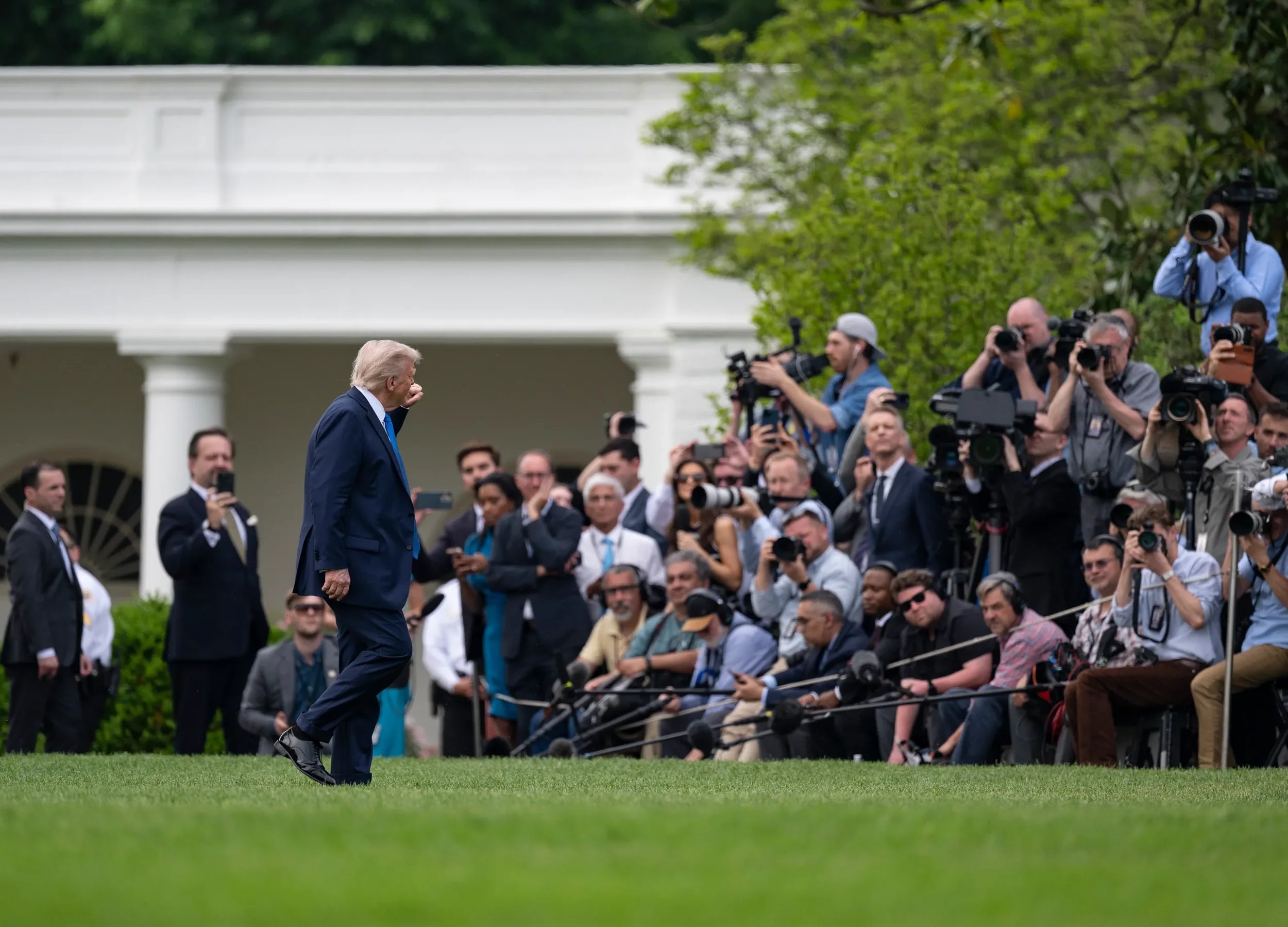Former President Donald Trump walking on the lawn with many journalists and photographers capturing his movement.