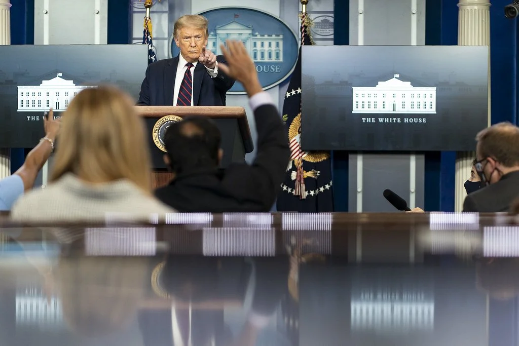 Donald Trump at the White House press briefing room, pointing forward, with audience members raising their hands, and two screens displaying the White House logo.