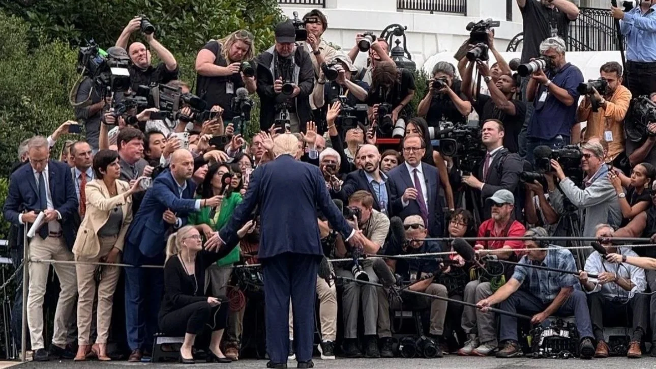 A person in a suit addresses a large crowd of reporters and photographers with cameras and microphones during a press conference outdoors in front of a white building.