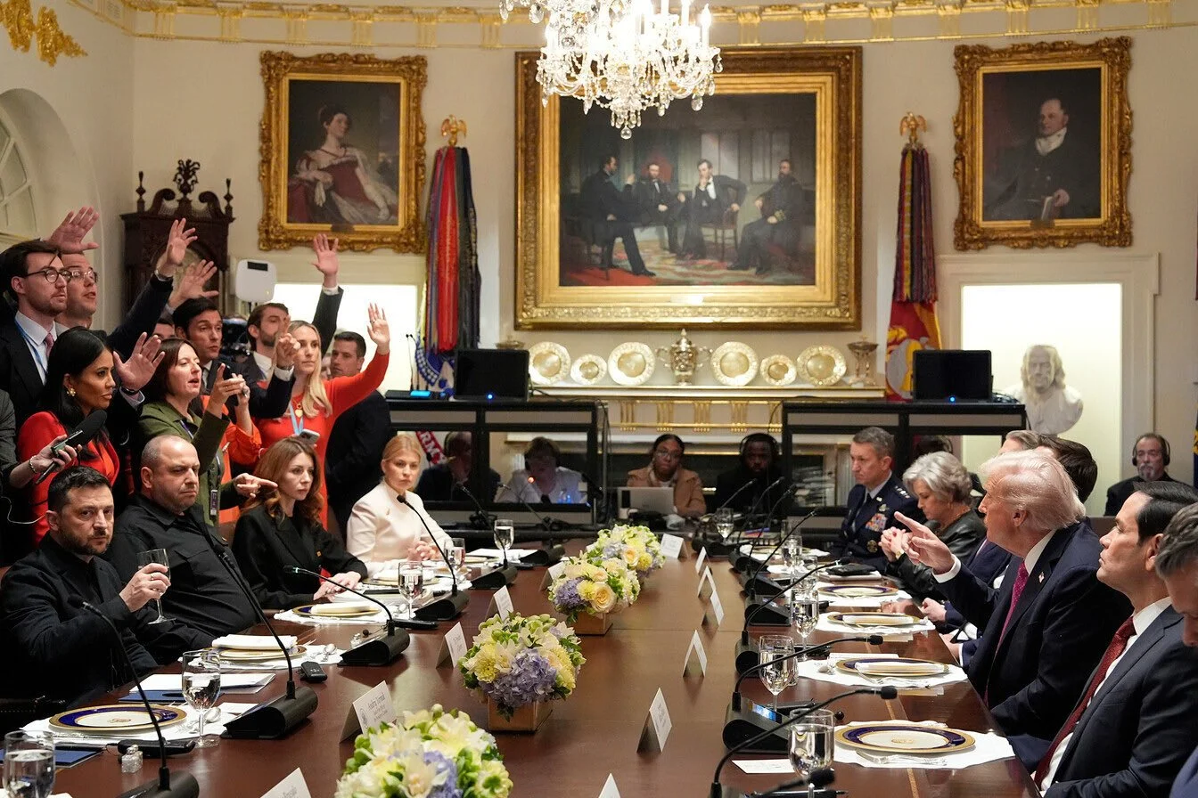 A formal meeting with world leaders at a long conference table decorated with flowers. Several people are raising their hands, and some are taking notes or looking at documents. The room has portraits and flags in the background.