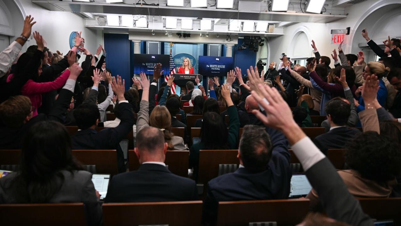 Press briefing in a government room with President at the podium, audiences raising hands, multiple screens behind showing White House logo.