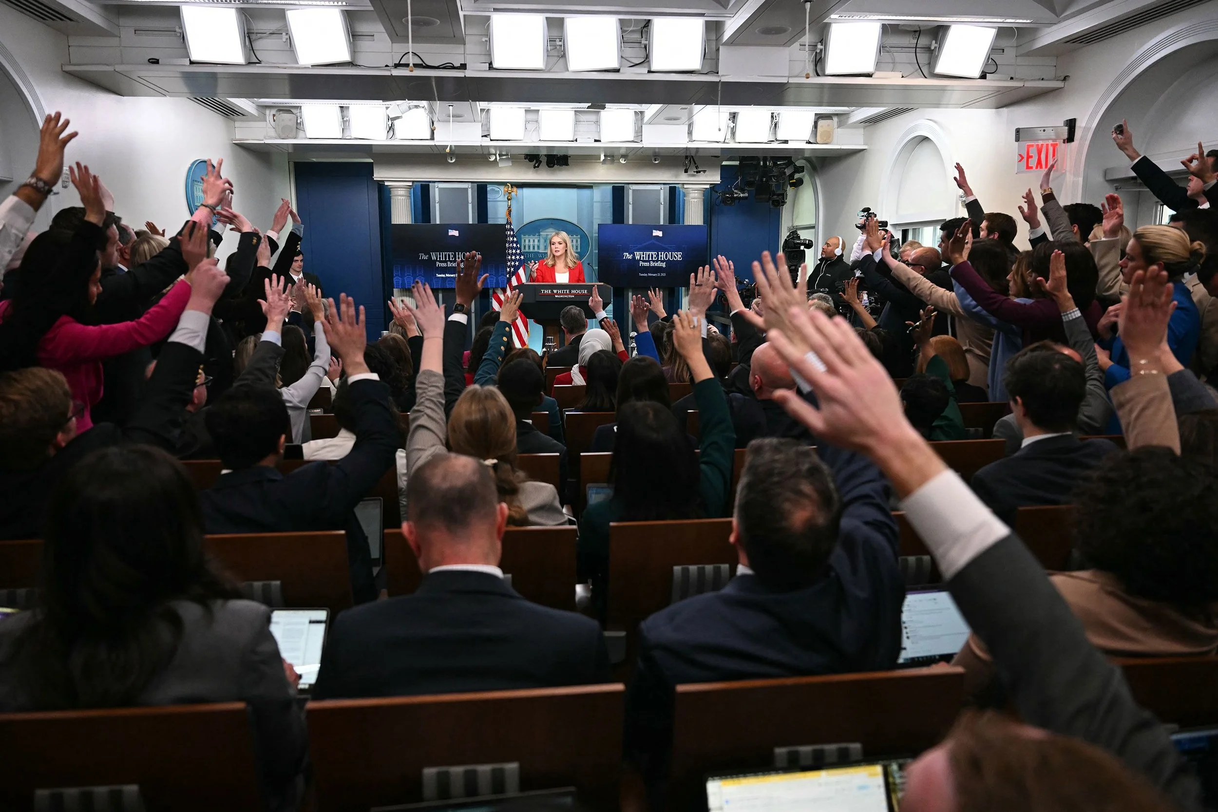 A woman at a podium giving a press briefing at the White House, with multiple screens behind her displaying the White House logo. The room is filled with people, many raising their hands, some taking notes on laptops, during a question-and-answer session.