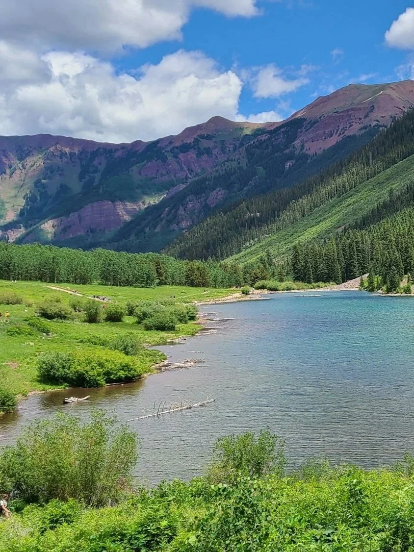 A scenic view of a river flanked by green grass and trees, with mountains covered in forest and purple vegetation in the background, under a partly cloudy blue sky in Colorado.