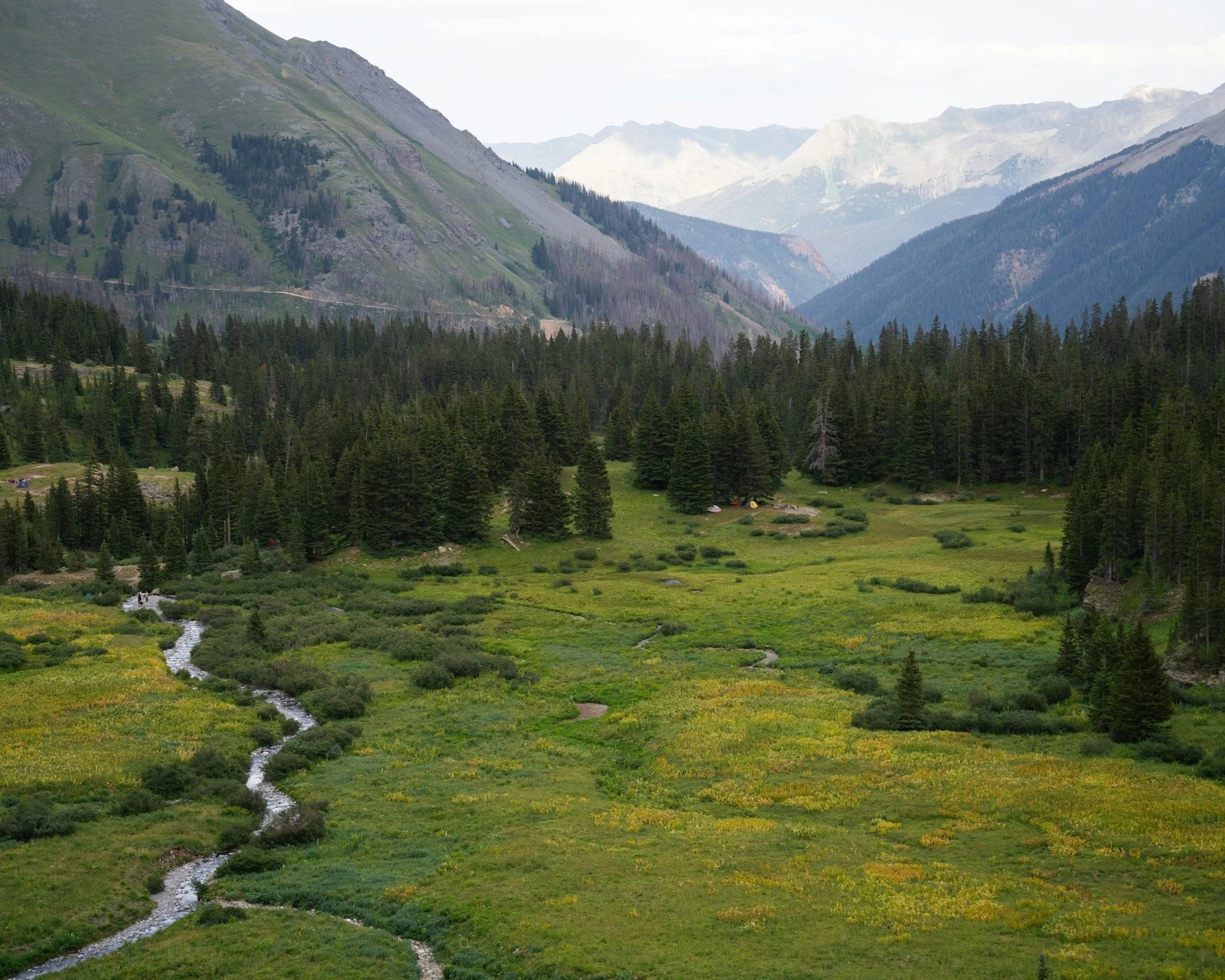 A scenic view of lush green mountains in Colorado with a small stream flowing through green meadows in the foreground, and taller mountains with tree-covered slopes in the background.