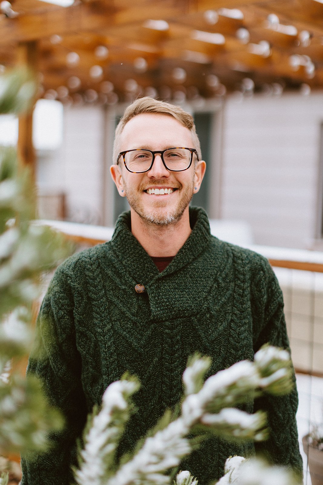 Matthew Eland with glasses and earrings, wearing a dark green cable-knit sweater, standing outdoors in front of a house with a wooden pergola.