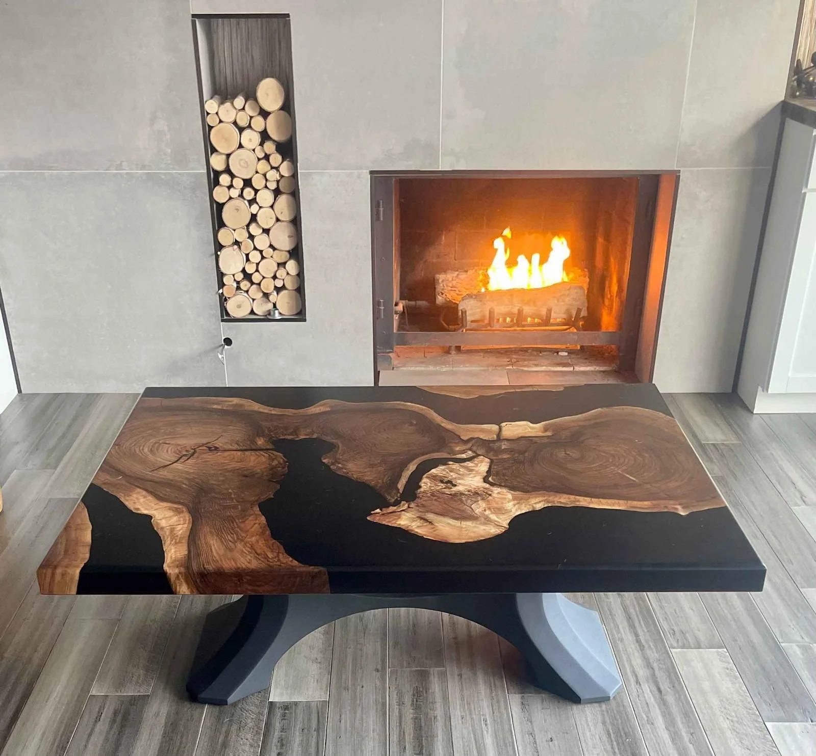 A rectangular, black and brown wooden table with a natural, irregular wood grain pattern in the center, and a metal base in a contemporary design. Behind the table, there is a fireplace with a fire burning and a wood storage niche filled with cut logs to the left.