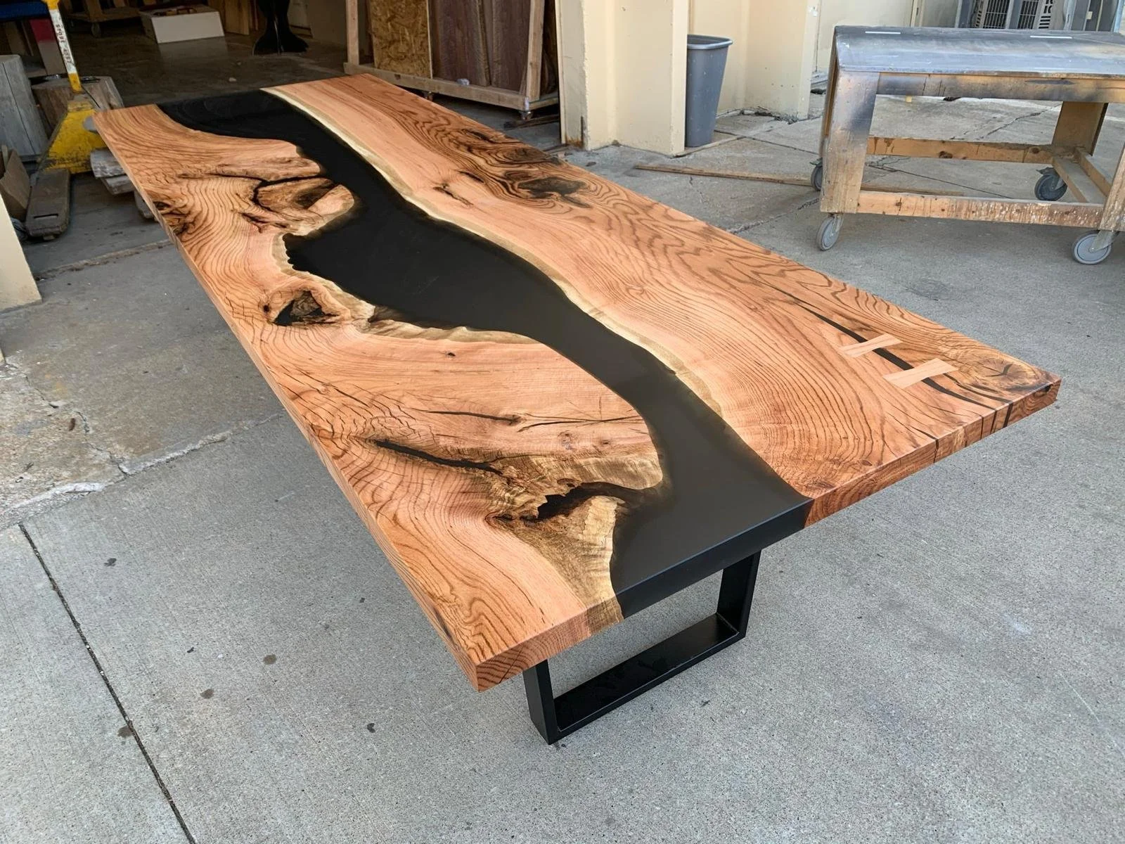 A rectangular wooden table with a natural edge and a black resin river design running through the center, placed outdoors on a concrete surface.